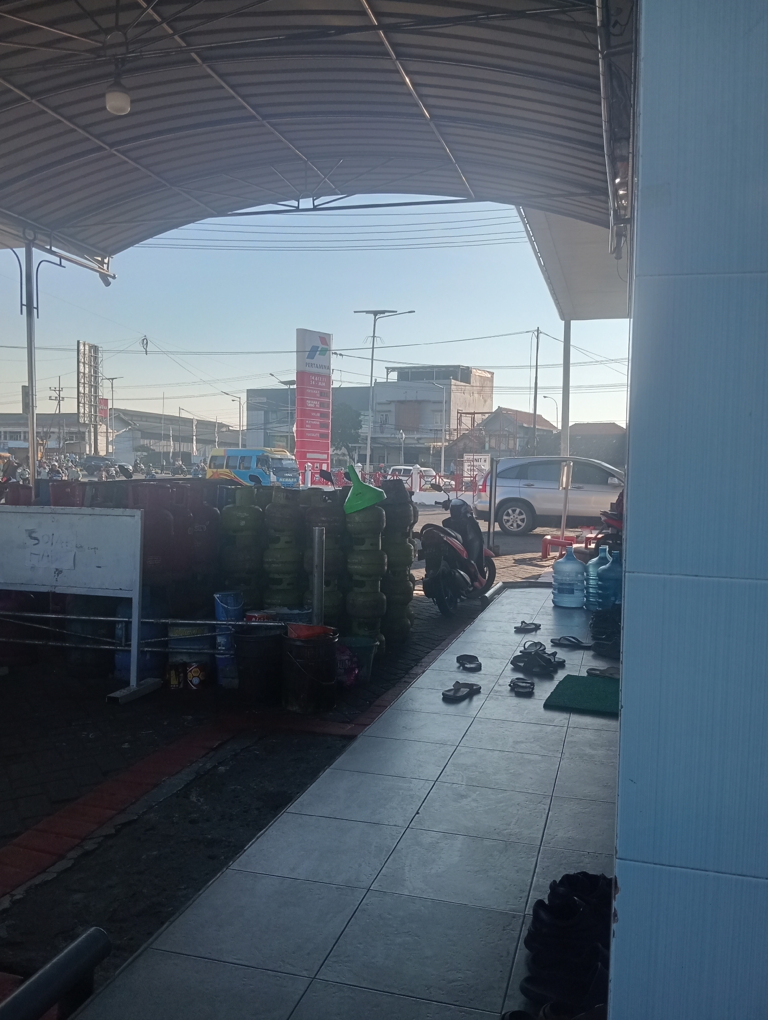 The image depicts an outdoor scene in Sawotratap, Indonesia, likely a gas station or a similar commercial area. The foreground shows a tiled walkway with numerous pairs of sandals and shoes scattered around. To the left, a large quantity of green propane tanks are stacked, suggesting a commercial establishment that sells or distributes them. A white sign with some writing is also visible to the left of the propane tanks. Beyond this, a road is visible with several motorcycles and a few cars, including a silver SUV, and a blue and yellow bus. A large red "PERTAMINA" sign, a prominent Indonesian state-owned oil and natural gas company, stands in the background, confirming the location as a fuel station. The sky is clear and bright, indicating daytime. The overall environment appears to be a bustling commercial hub.