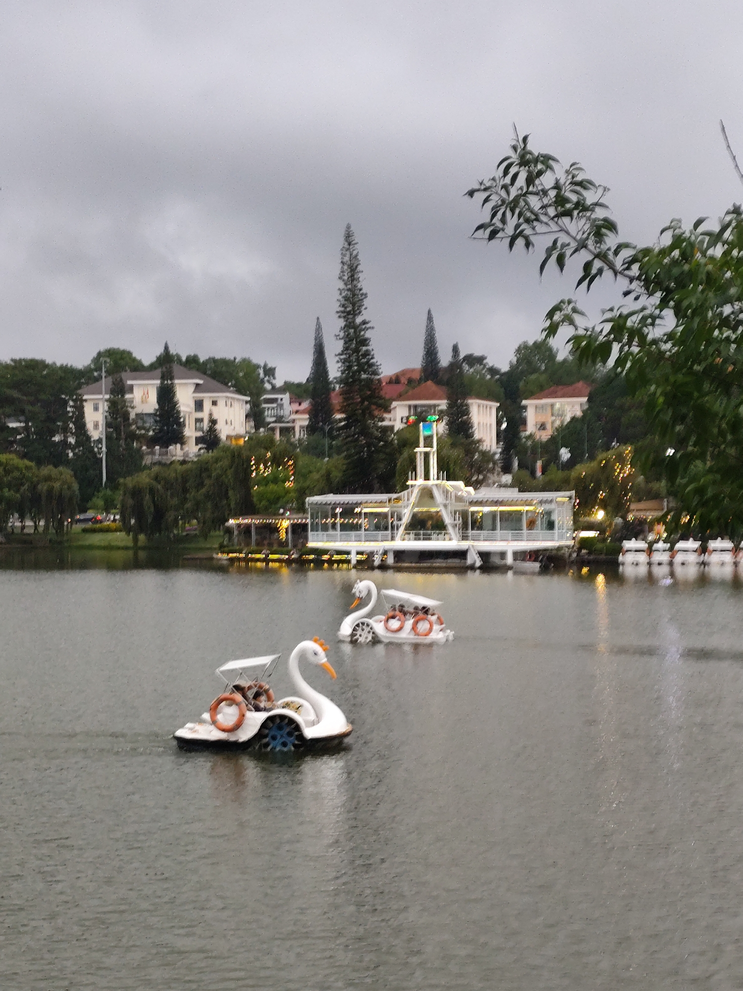 The photograph depicts a cloudy day scene at Ho Xuan Huong Lake in Da Lat, Vietnam. Two swan-shaped paddle boats, one occupied, are visible on the water. The shore is lined with trees, buildings, and a multi-level white structure adorned with lights. The overall lighting is dim due to the overcast sky.