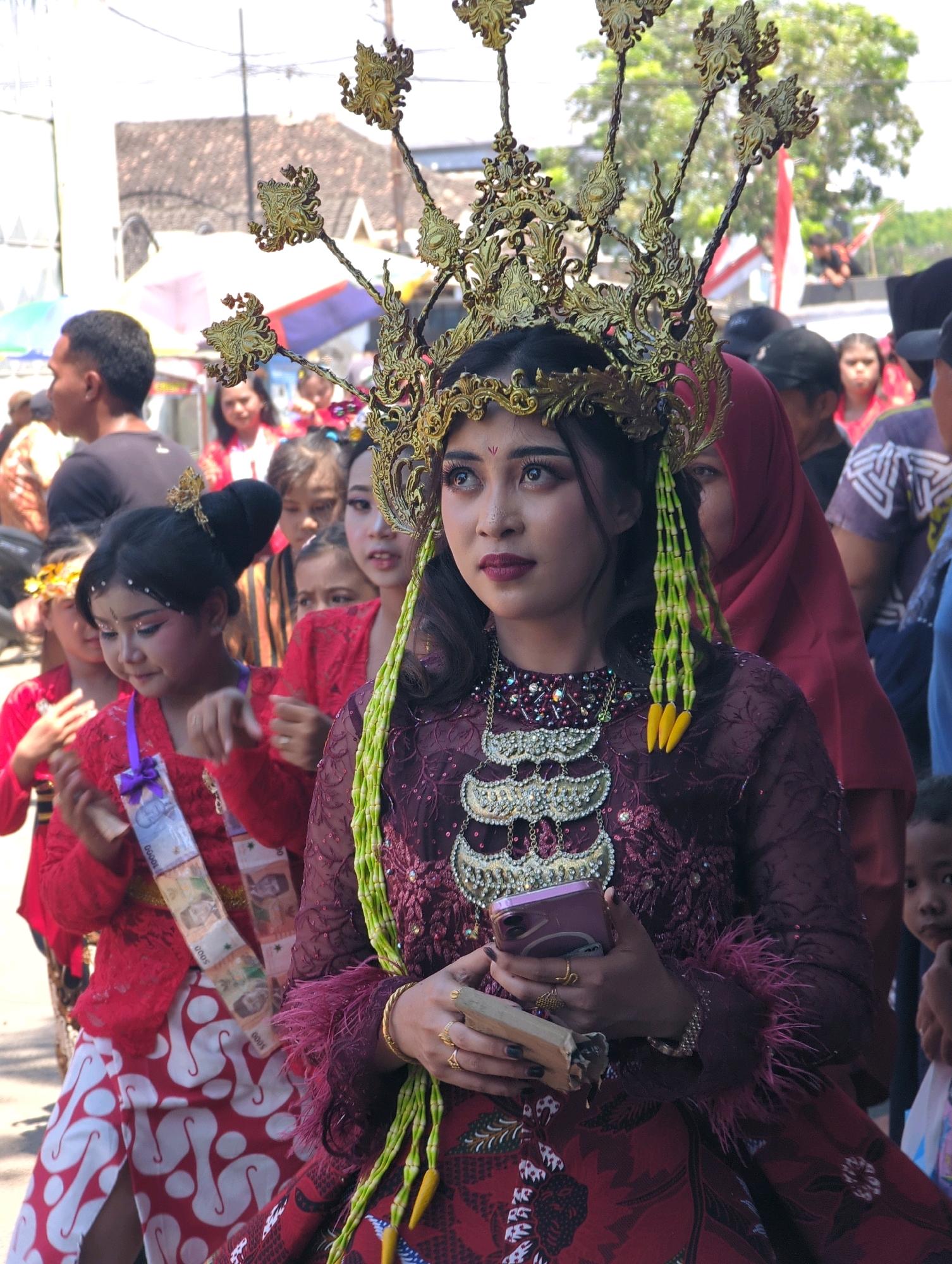 In Pasuruan, Indonesia, a young woman adorned in elaborate traditional attire, including a gold headdress with ornate detailing and a deep red, intricately embellished dress, stands centrally. Her makeup is dramatic, with bold eyeliner and lipstick. She holds a smartphone and a rectangular object in her hands, looking upwards and to the side. To her left, several children in similar red outfits, one with a sash featuring banknotes, also appear to be part of the event. Behind them, a crowd of spectators and participants, some wearing headscarves and traditional Indonesian clothing, are visible. The background suggests an outdoor setting in a populated area, with buildings and trees under a bright sky. The overall scene depicts a cultural event or procession.