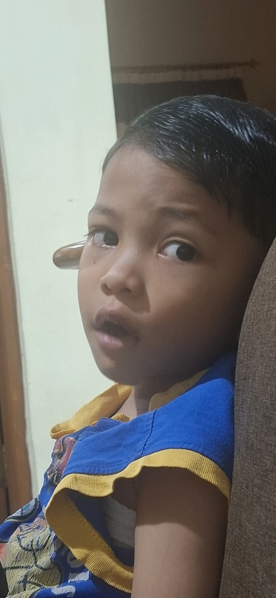 A medium shot portrait shows a young child with dark hair and brown eyes looking towards the viewer's left. The child is wearing a blue t-shirt with yellow trim. The background is a light-colored wall. The image appears to be taken indoors. The overall scene is dimly lit, with some shadowing. The child's left eye appears to be partially obscured by what could be a reflection or part of the environment. The child's mouth is slightly open, and their expression is neutral. The child's head is tilted slightly, and they are positioned on the right side of the frame.