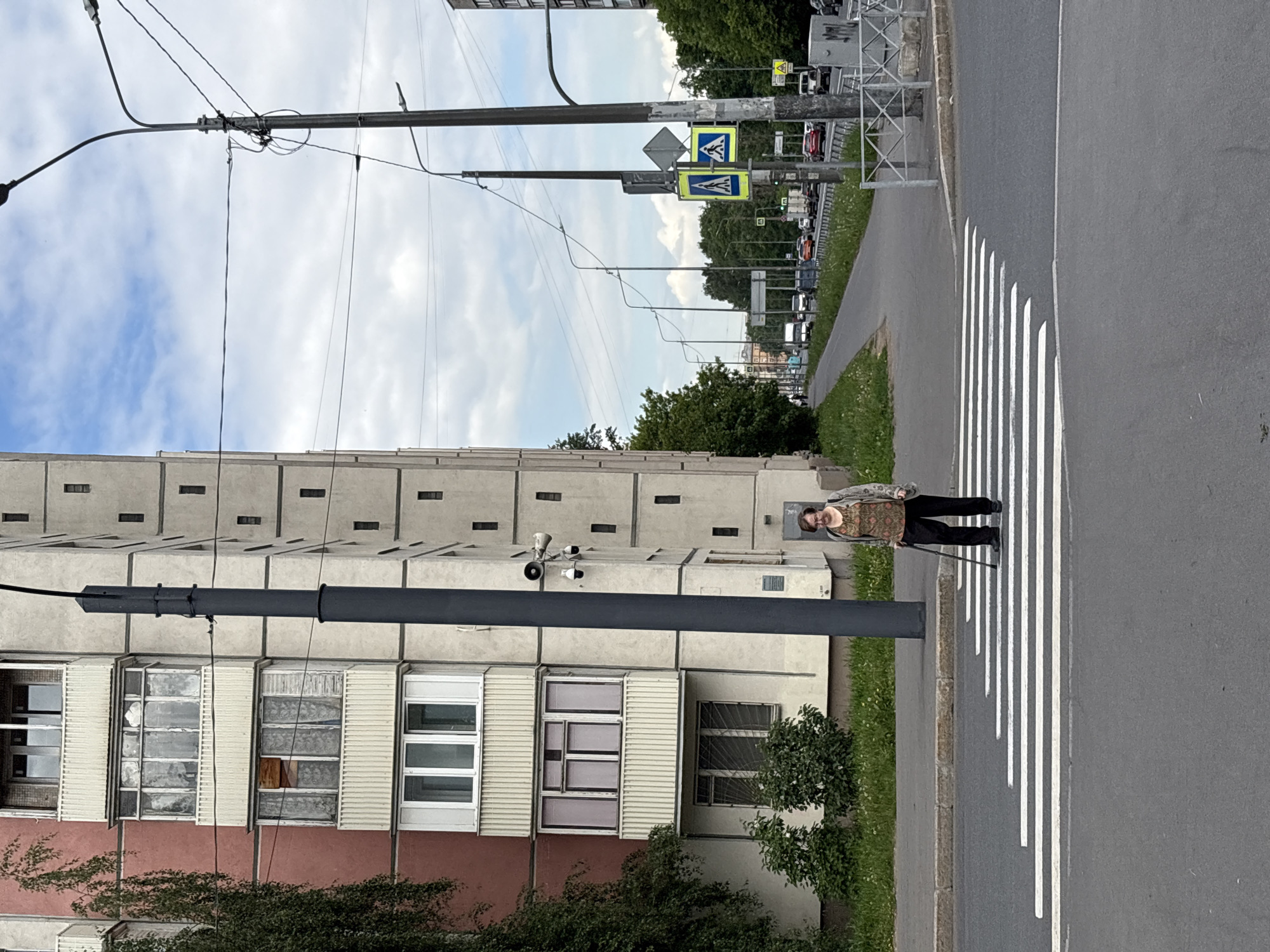 This rotated image captures an urban street scene in Saint Petersburg, Russia, on an overcast but bright day.
The primary subject is an elderly person, likely a woman, positioned on a white zebra crossing. She is wearing dark trousers and a patterned top, and she supports herself with a cane.
To her left (when correctly oriented), a tall, multi-story residential building dominates the frame. Its upper floors feature a distinctive light grey facade with repetitive small, rectangular indentations, while the lower floors display a variety of windows, some appearing aged. The base of the building is a reddish-brown color. Two security cameras are visibly mounted on the building's facade, along with a utility pole.
On the right side of the street, beyond the pedestrian crossing, a grey paved sidewalk is bordered by a strip of green grass. Further down the road, two blue and yellow pedestrian crossing signs are attached to a pole, marking the crossing. The background reveals a street lined with trees, distant buildings, and what appear to be parked cars, indicating an active urban environment. Numerous overhead power lines and streetlights crisscross the sky above the street.
The person appears to be paused on the crossing, possibly in the process of traversing the street. No discernible text is visible other than the universal pedestrian symbols on the traffic signs.