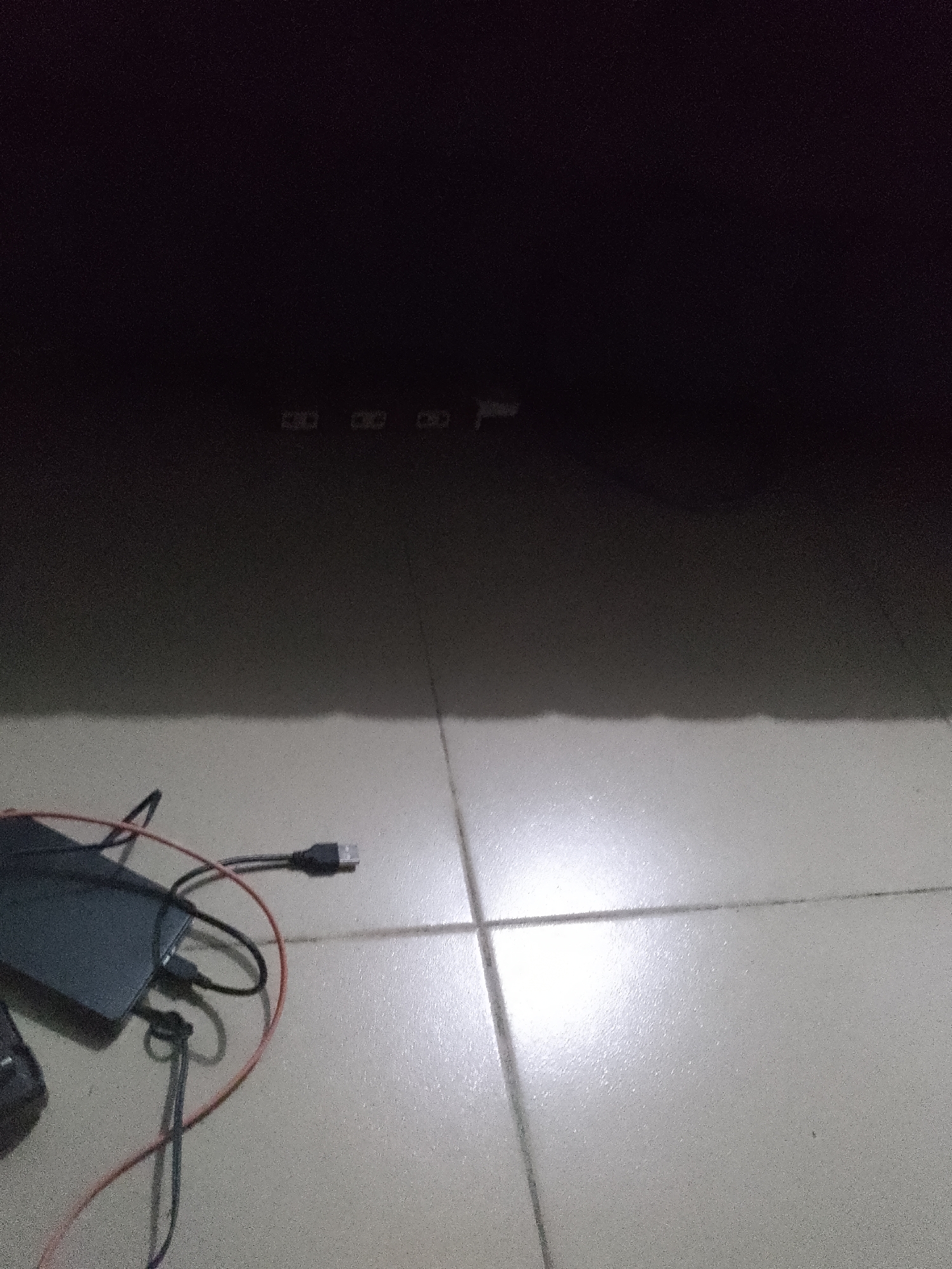 An overhead, low-light photograph shows a tiled floor in Jauro Yinu, Nigeria. In the lower left quadrant, a black external hard drive is visible, connected to a black USB cable and a red cable. The hard drive is situated on the floor, casting a shadow. In the upper portion of the image, a dark, indistinct shape suggests a piece of furniture. A shadow cast by this furniture stretches across the tiled floor, creating a gradient of light and dark. The tiles are a light color, and the grout lines are visible.