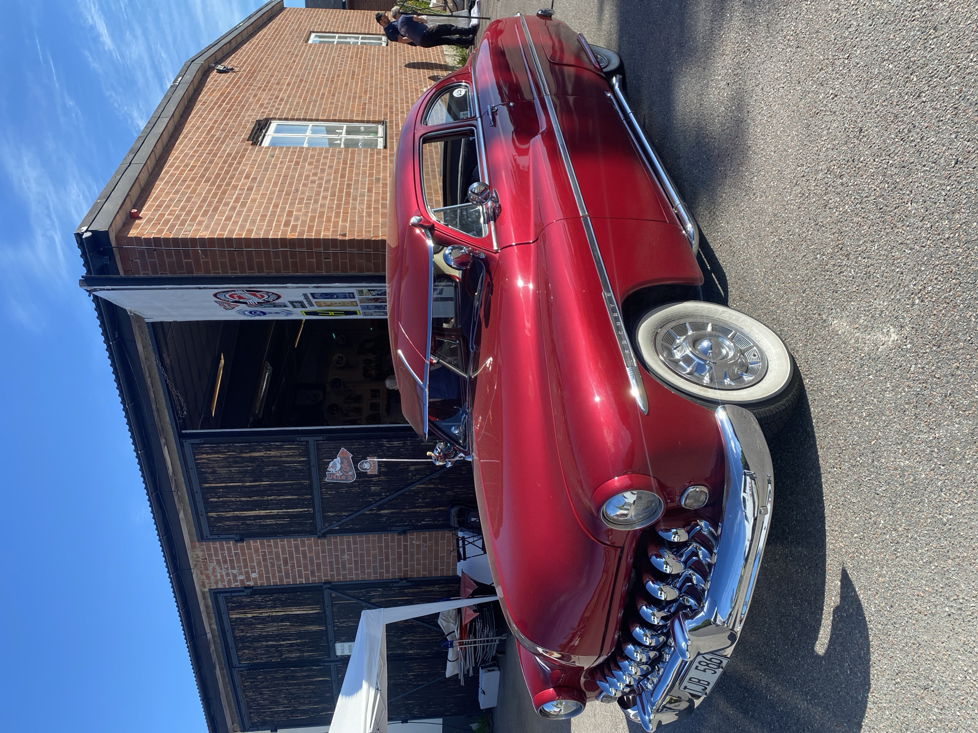 A shiny, red, vintage Mercury coupe is parked on an asphalt surface in front of a brick building with large wooden double doors. The building appears to be a garage or workshop, with a corrugated metal roof. A white canopy is set up to the left of the building, and various items, possibly for sale or display, are underneath it. Two people, dressed in dark clothing, stand near the rear of the building, one interacting with something on the ground. The sky is blue with scattered white clouds, indicating a daytime scene. The license plate on the car reads "TJB 586". The overall setting suggests a car show, vintage car event, or an open house at a vehicle-related business in Skyttorp, Sweden.