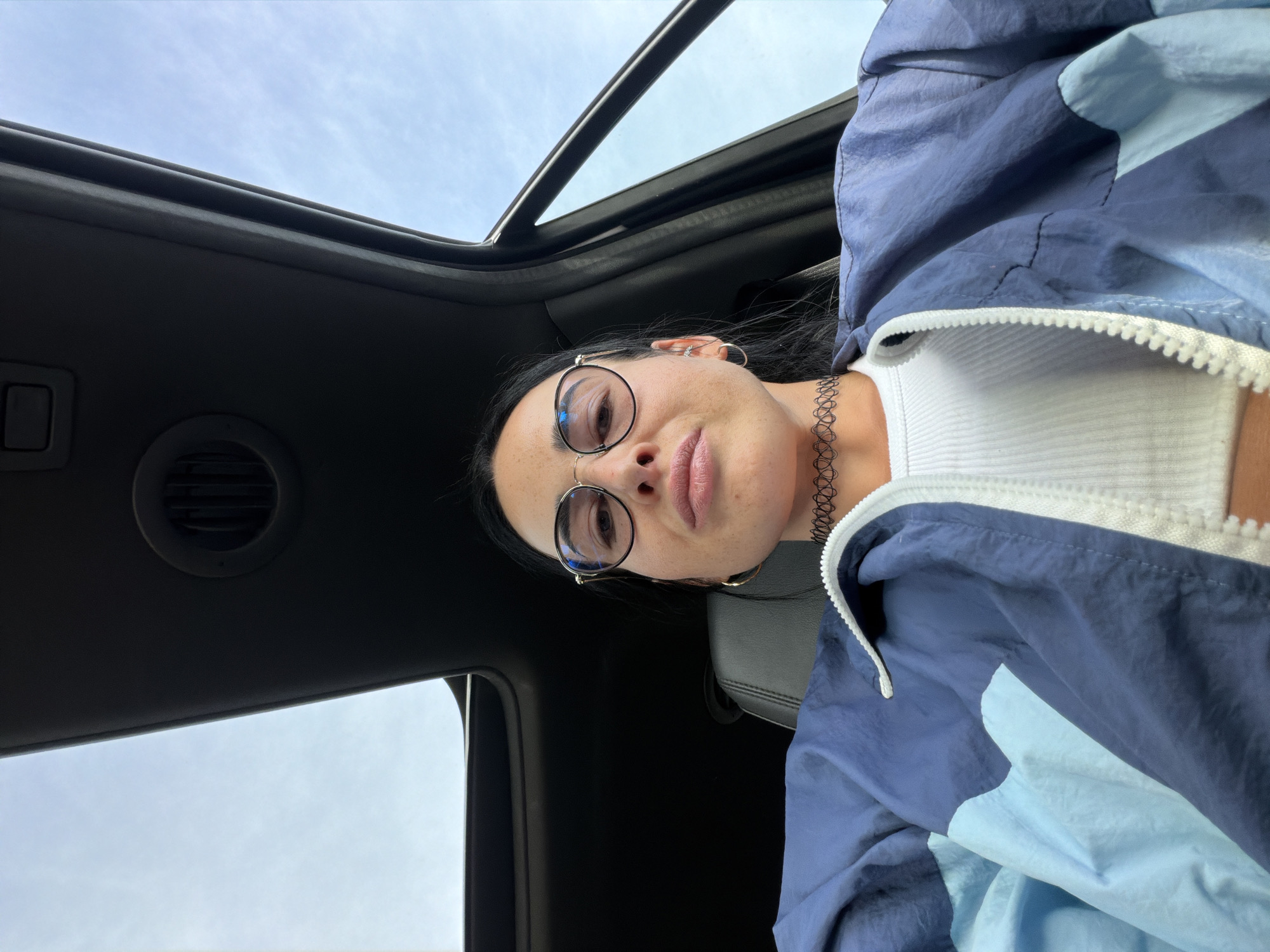 A woman is taking a selfie inside a vehicle. She is wearing a white ribbed tank top, a blue and light blue color-blocked windbreaker, round glasses, and a black choker necklace. Her face is centered in the frame, with her head tilted slightly. The sunroof of the vehicle is open, revealing a cloudy sky. The interior of the car is dark, with a visible air vent on the ceiling. The setting is the city of Sholokovo, Russia.