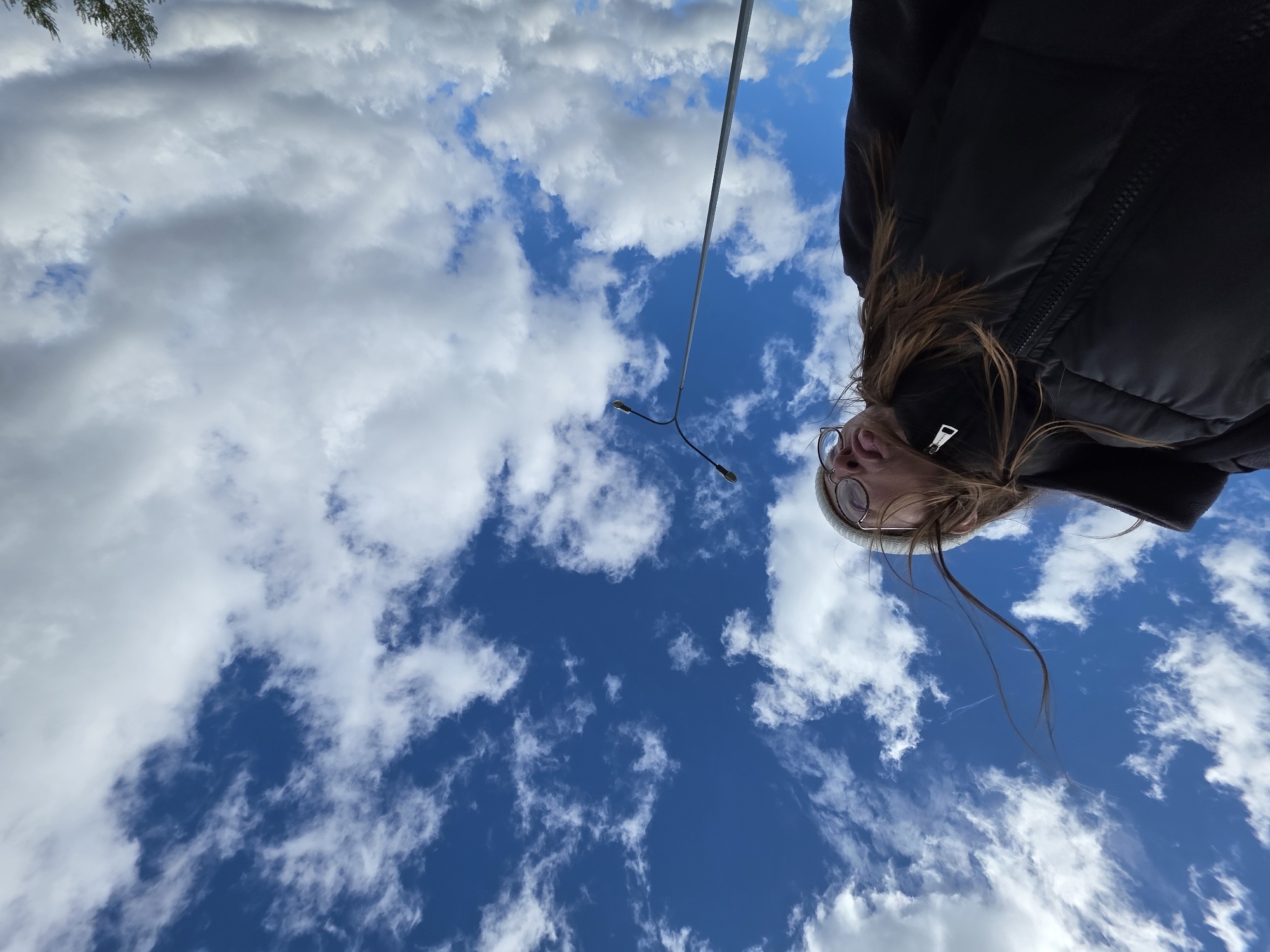 A low-angle shot depicts a person wearing a black puffer jacket and glasses, with their face tilted upwards towards a sky filled with white clouds and patches of blue. A street lamp is visible in the mid-ground, positioned between the person and the sky. Based on metadata (not visible in the image but previously available), this photo was taken in Chaikovsky, Russia.