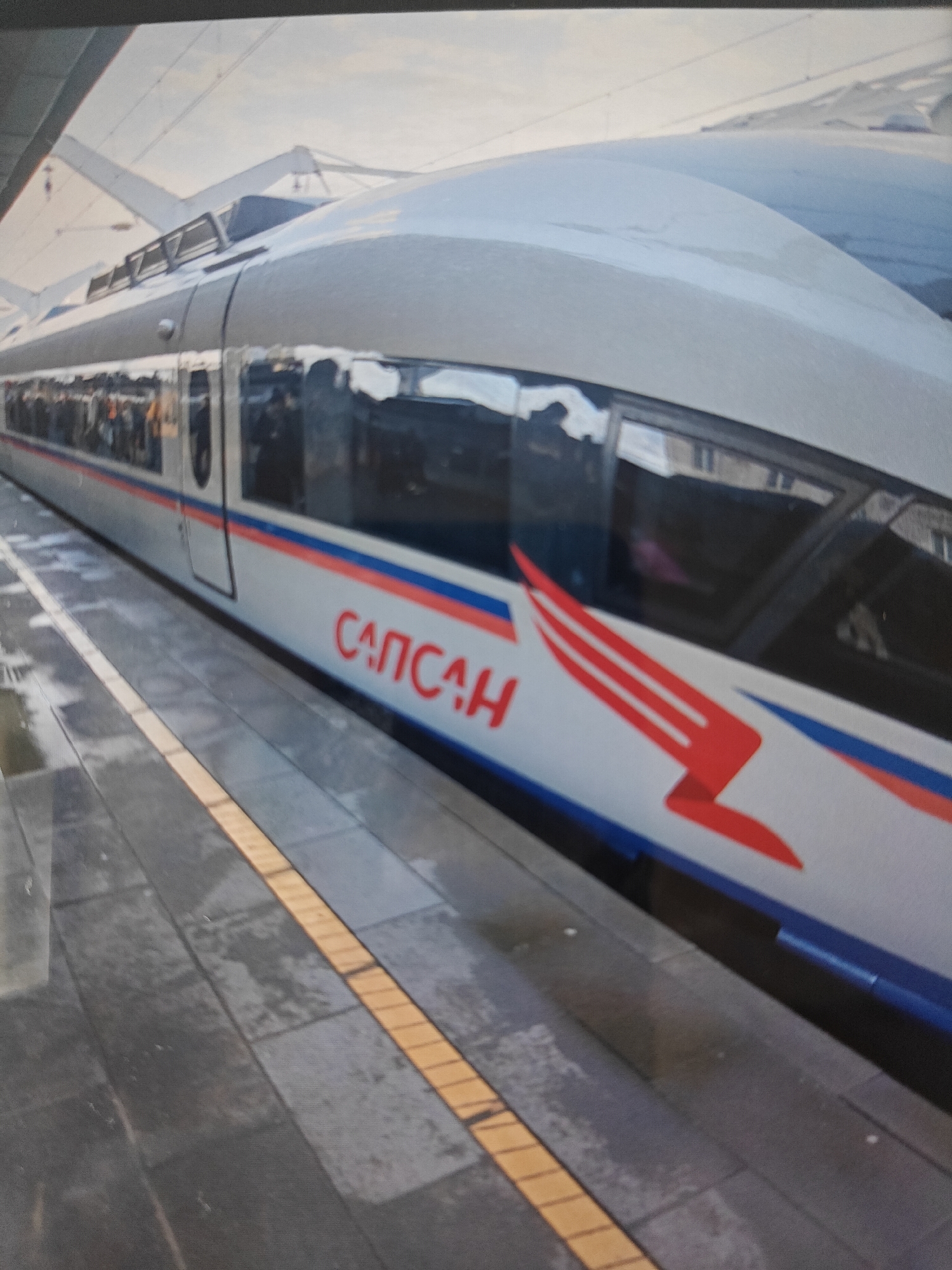 A high-speed train identified by the Cyrillic text "САПСАН" (Sapsan) is stationary at a platform. The train is white with red and blue accents, featuring a red stylized wing graphic on its side. The platform consists of gray tiles with a yellow tactile warning strip running along the edge. The train's windows reflect the surrounding environment, showing hints of overhead structures and possibly other trains. The sky appears overcast. The scene is set in Pushkino, Russia.