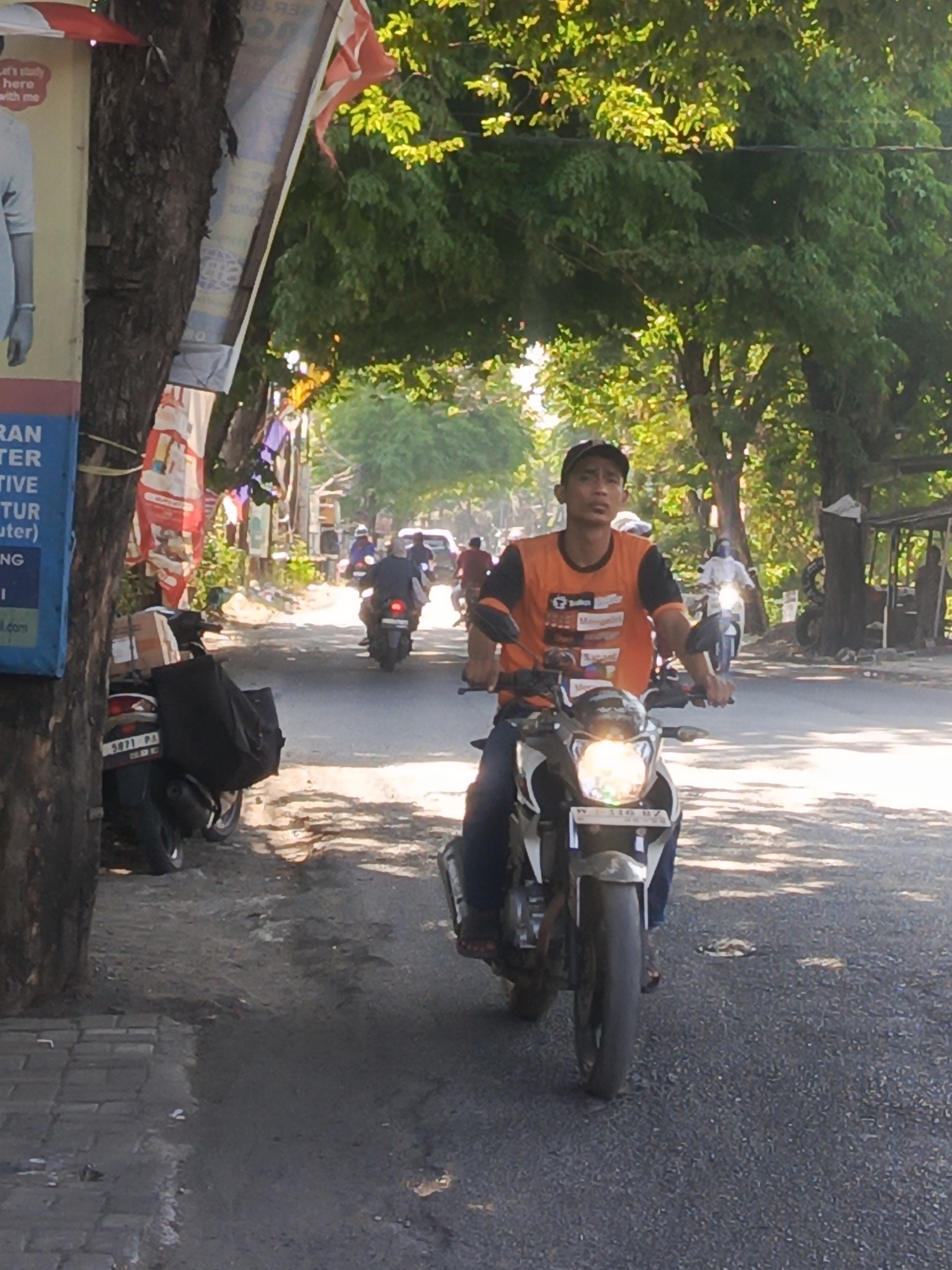 A young man wearing an orange t-shirt with black sleeves and a black baseball cap rides a white motorcycle on a paved road in Sidowungu, Indonesia. He is looking forward with a neutral expression. The motorcycle's headlight is on. Behind him, other motorcycles and a car are visible on the road. The scene is bordered by trees on the left and right, with sunlight filtering through the leaves. A large tree trunk is prominent on the left side of the image, with posters attached to it. A parked motorcycle with luggage is visible to the left of the main subject. The road is asphalt and appears to be in fair condition, with some debris and patches of dirt. The time of day appears to be morning or afternoon, given the sunlight.