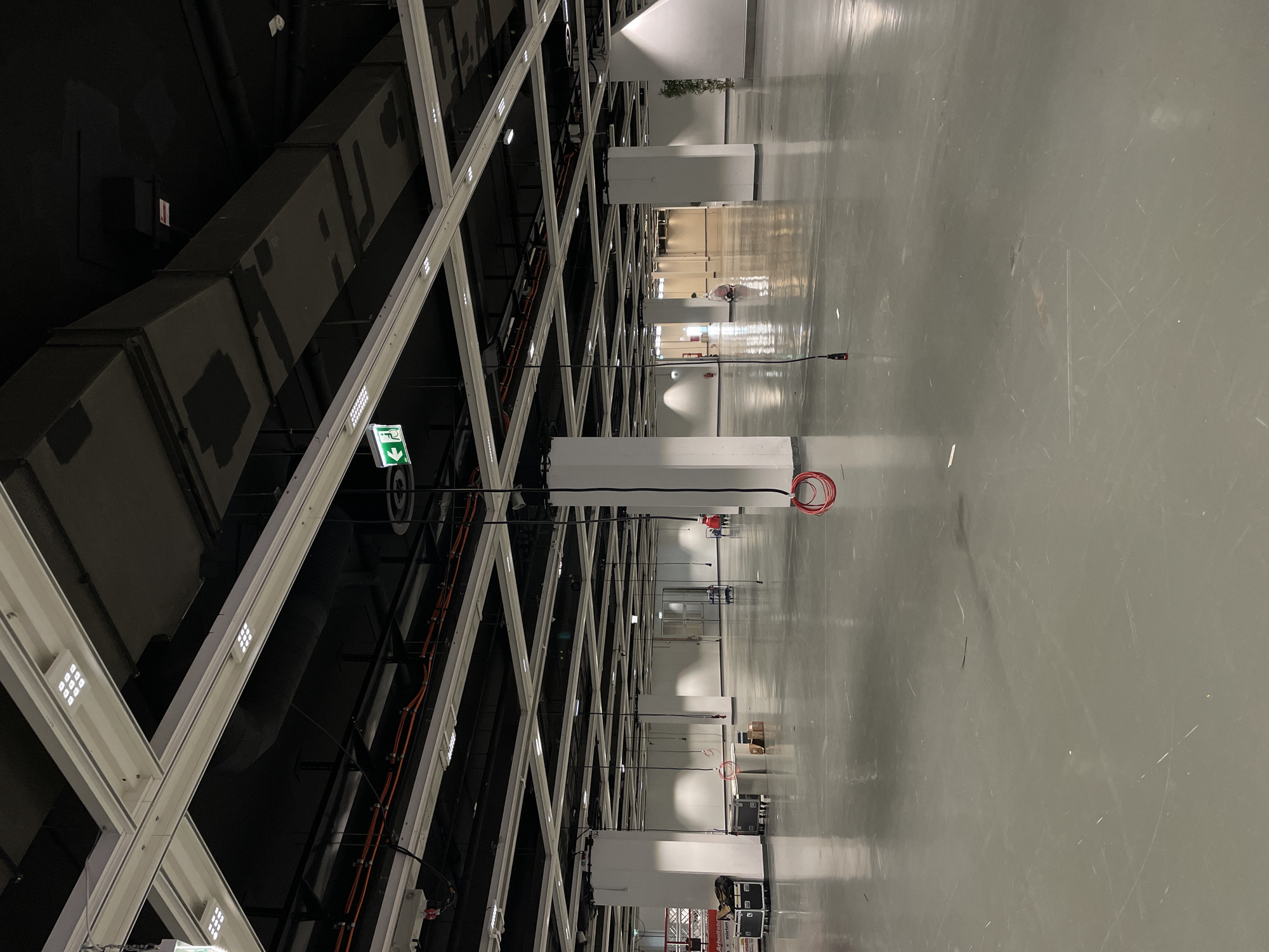 The image depicts a large, modern interior space, possibly a convention center or exhibition hall, characterized by a grid-like ceiling structure of white beams and black ductwork. Rows of white, rectangular fixtures descend from the ceiling, spaced evenly along the length of the room. The floor is a reflective grey surface, likely polished concrete, with visible scuff marks and streaks. In the distance, human figures are partially visible, along with various objects and structures suggesting an active, functional space. A green emergency exit sign is visible on one of the ceiling beams. Orange electrical conduits are visible running along the ceiling beams. The location within a city of Unknown, Unknown cannot be determined from the image.