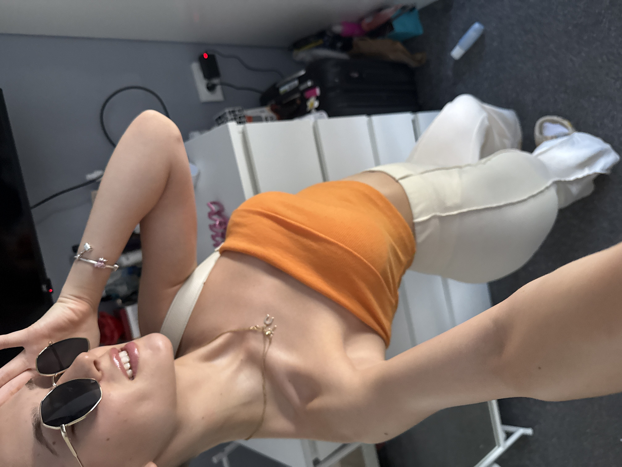 A young woman is shown from the chest up, posing for a selfie in what appears to be a bedroom. She is wearing large, gold-rimmed sunglasses and an orange, off-the-shoulder top. A delicate gold necklace with a small pendant is visible on her collarbone. Her left hand is raised, touching the side of her head. The background reveals a white dresser with drawers, a black suitcase, and other indistinct items, suggesting a domestic setting. The lighting indicates natural light coming from a window to the woman's left. The image does not contain any elements that would allow for its location to be specified as Sofia, Bulgaria.