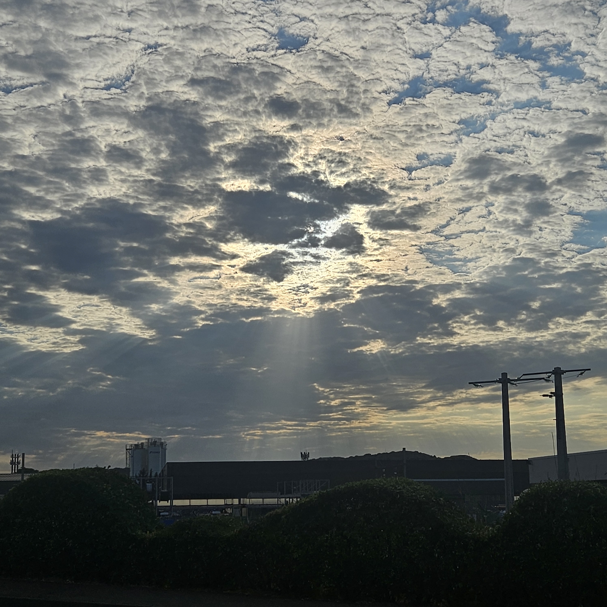 The image shows a sky filled with cumulus clouds, with the sun partially obscured by them, creating crepuscular rays that pierce through the clouds. In the foreground, a row of dark green bushes lines the bottom of the frame. Behind the bushes, a dark, low-lying structure, possibly a building or industrial facility, stretches horizontally across the middle ground. To the left of this structure, a tall industrial silo with visible piping is present. To the right of the structure, two utility poles with wires are visible. The overall scene suggests an industrial or semi-urban environment under a dramatic sky. The location is identified as the city of Narbonne, France, implying the structures and landscape are specific to that area. There are no people or animals visible in the image.