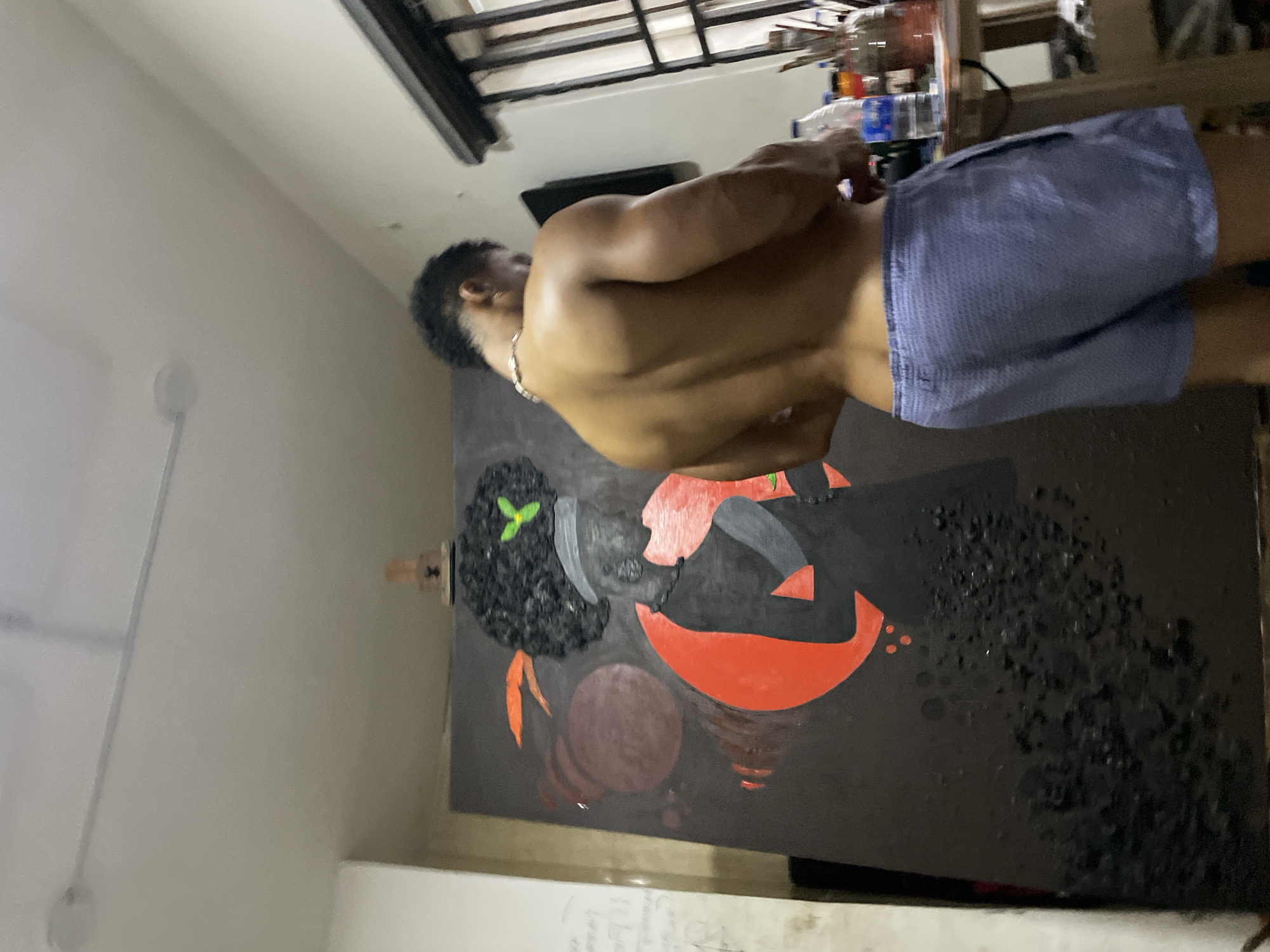 A person is standing in a room in Ado, Nigeria, facing a wall with a painting. The painting depicts a figure with dark hair, an orange design, and other abstract elements in red, black, and other colors. The person has dark hair and is wearing light blue shorts. The room has a white wall, a metal-framed ceiling fixture, and various items on a shelf or counter in the background. The person's back is visible.