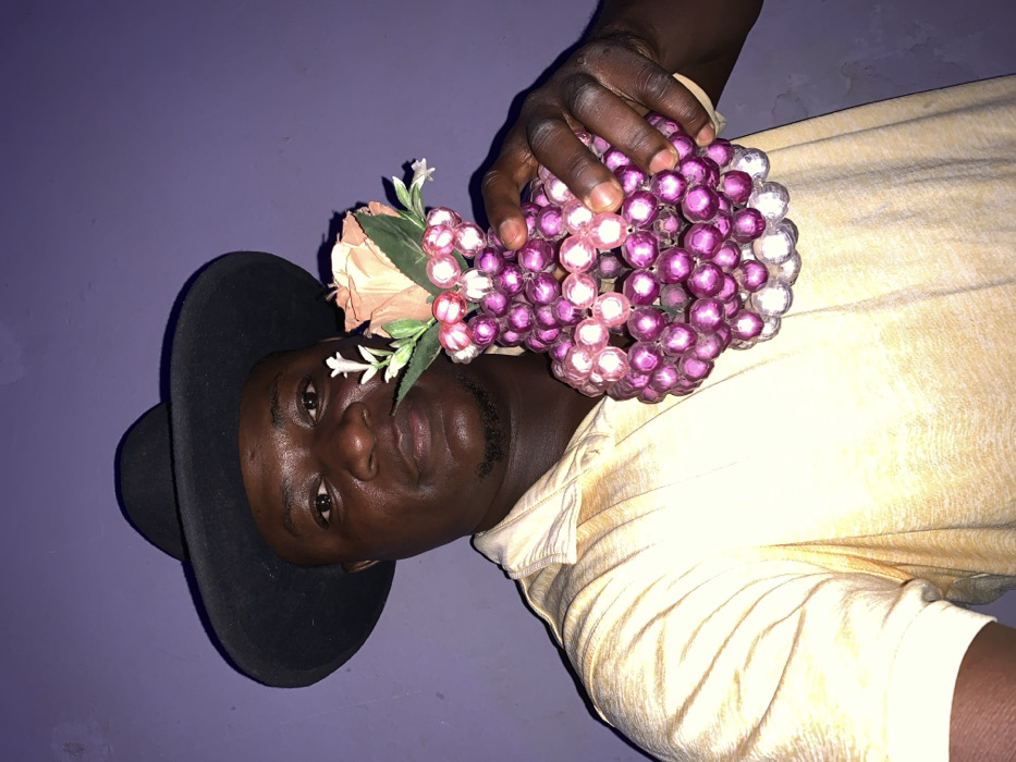 A man with dark skin and a black hat is seen lying on his side. He wears a pale yellow shirt and has a small beard. He holds a sphere made of pink and purple beads in his hand. The sphere also contains some artificial flowers with green leaves. The background is a solid purple color. The photograph is slightly angled, with the man's head near the bottom left corner.