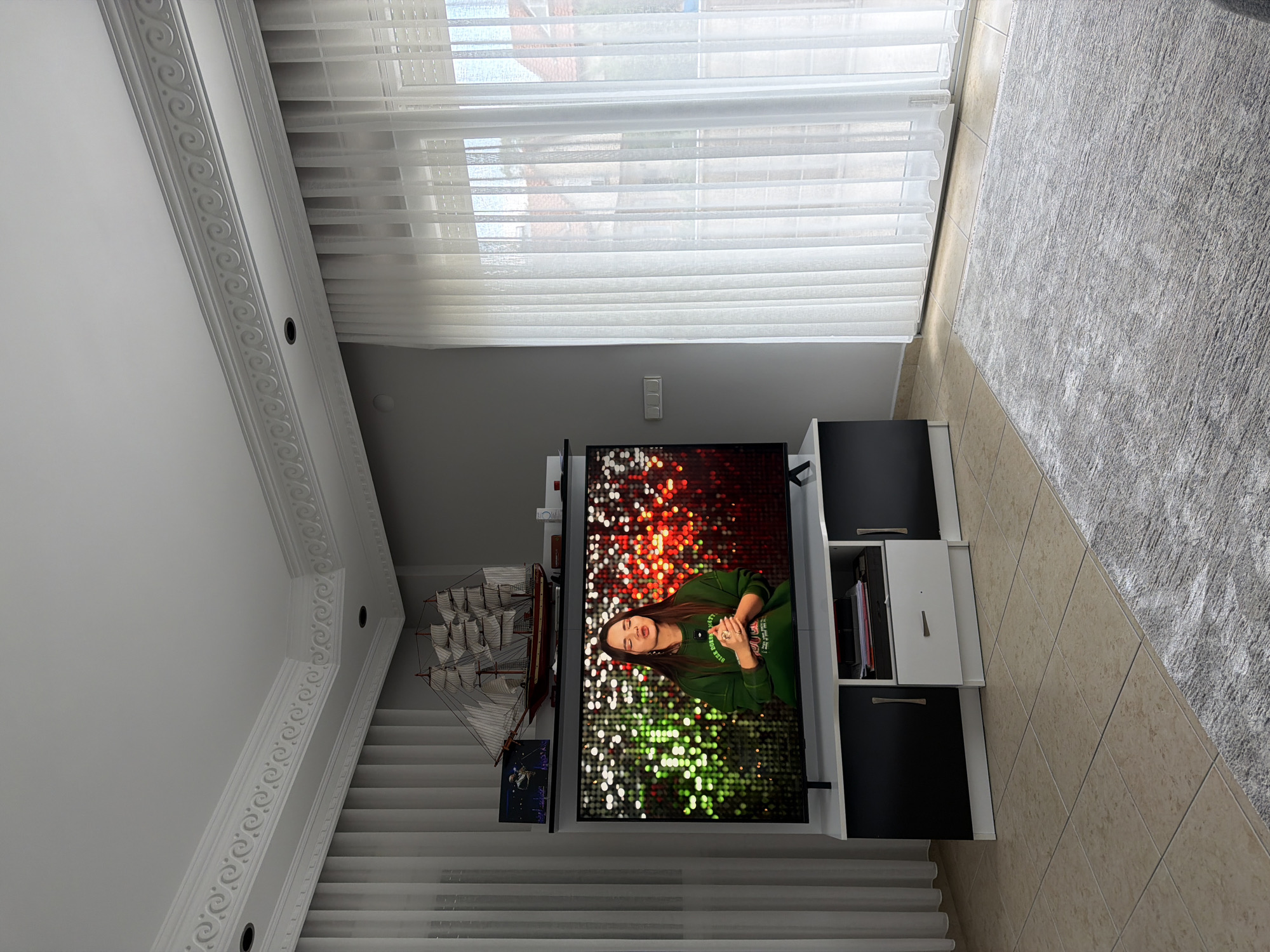 The image depicts an indoor living room scene. A large television screen displays a young woman wearing a green hoodie and speaking, with a colorful, bokeh background. To the left of the TV, a model ship is positioned on a shelf, alongside a framed picture. The television is situated on a black and white media console. The room features tiled flooring, a light-colored rug, and sheer vertical blinds covering a window or glass door. The walls are painted a neutral gray. The ceiling has decorative crown molding.