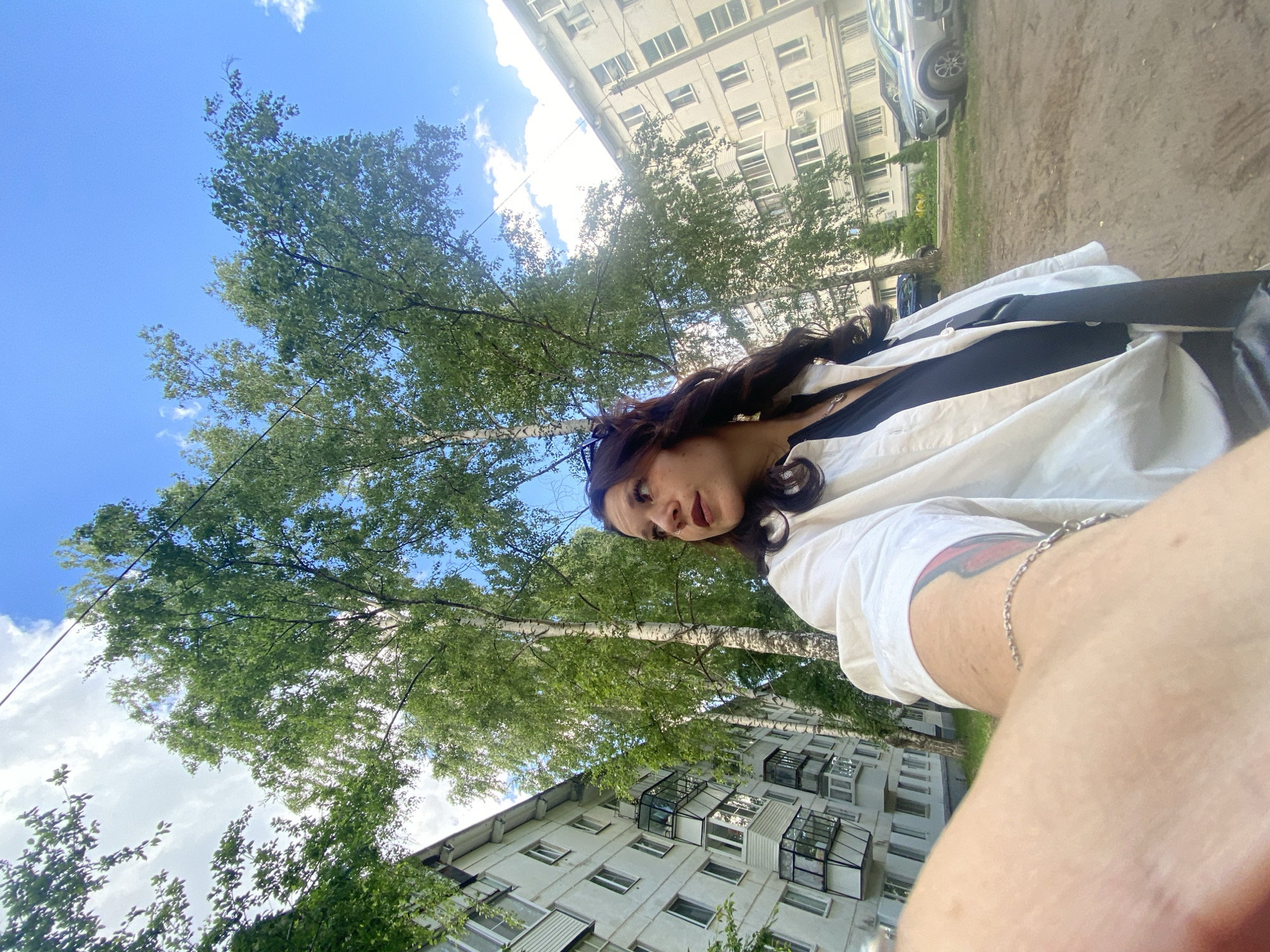 A first-person perspective selfie shows a young woman with long, dark, wavy hair and makeup, wearing a white unbuttoned shirt over a dark top and a silver chain necklace. She is looking to her right, with a faint smile. In the background, there are two multi-story apartment buildings, several tall trees with green foliage, and a blue sky with scattered clouds. Cars are parked in the dirt lot between the buildings and trees. The scene appears to be in a residential area of Chaikovsky, Russia, likely during daylight hours given the lighting. The overall impression is one of a casual outdoor setting.