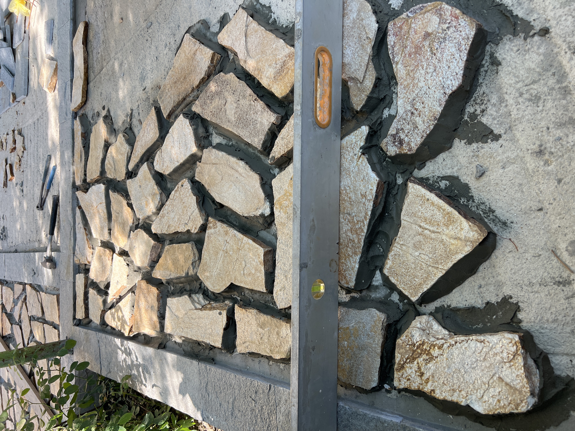 The image captures a section of landscaping work in progress in Menenkyovo, Bulgaria. The foreground shows a pathway being laid with irregular, flat stones set in mortar. A spirit level is positioned horizontally across the laid stones, indicating an effort to ensure a level surface. The stones are predominantly light brown and beige, with some showing hints of reddish-brown and white speckling. The mortar is dark gray and fills the gaps between the stones. In the upper portion of the image, more stones are visible, both laid and stacked, along with some gardening tools. To the left, lush green foliage suggests an outdoor setting, possibly a garden or park. The overall scene depicts manual construction and meticulous attention to detail in creating an outdoor pathway.