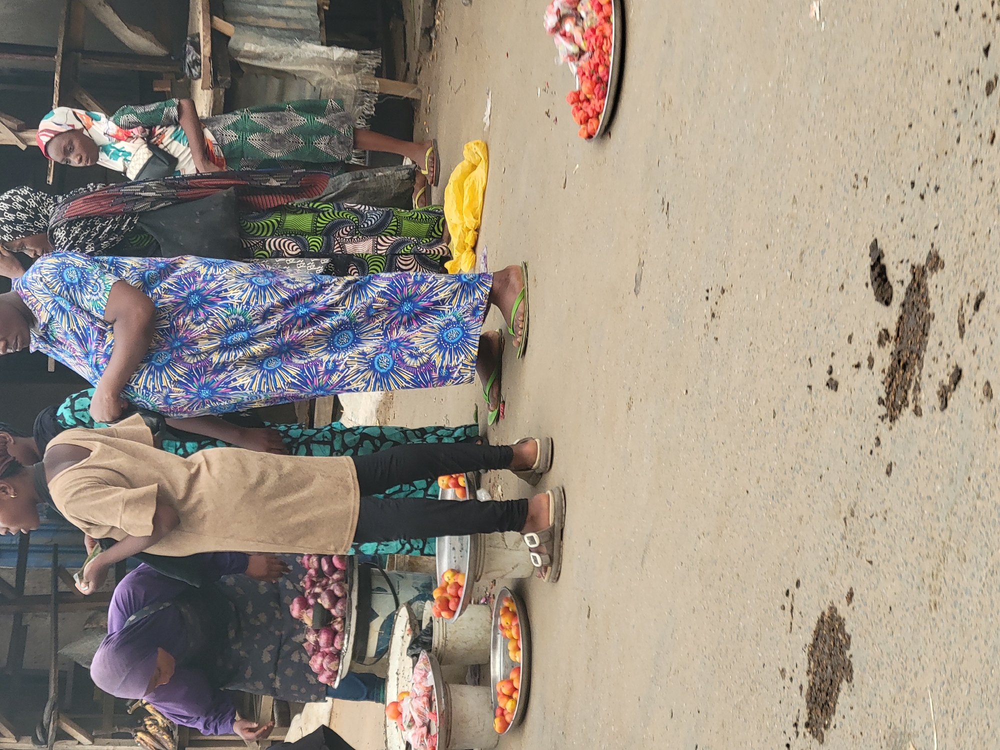 The image depicts an outdoor market scene in Abuja, Nigeria. Several women are visible, each standing behind a display of goods on metal trays or in baskets. The items for sale appear to be produce, including tomatoes, onions, and red peppers. The women are dressed in various traditional African garments. The setting is an urban environment, with what looks like buildings and structures in the background. The ground is a paved surface with some dark stains.