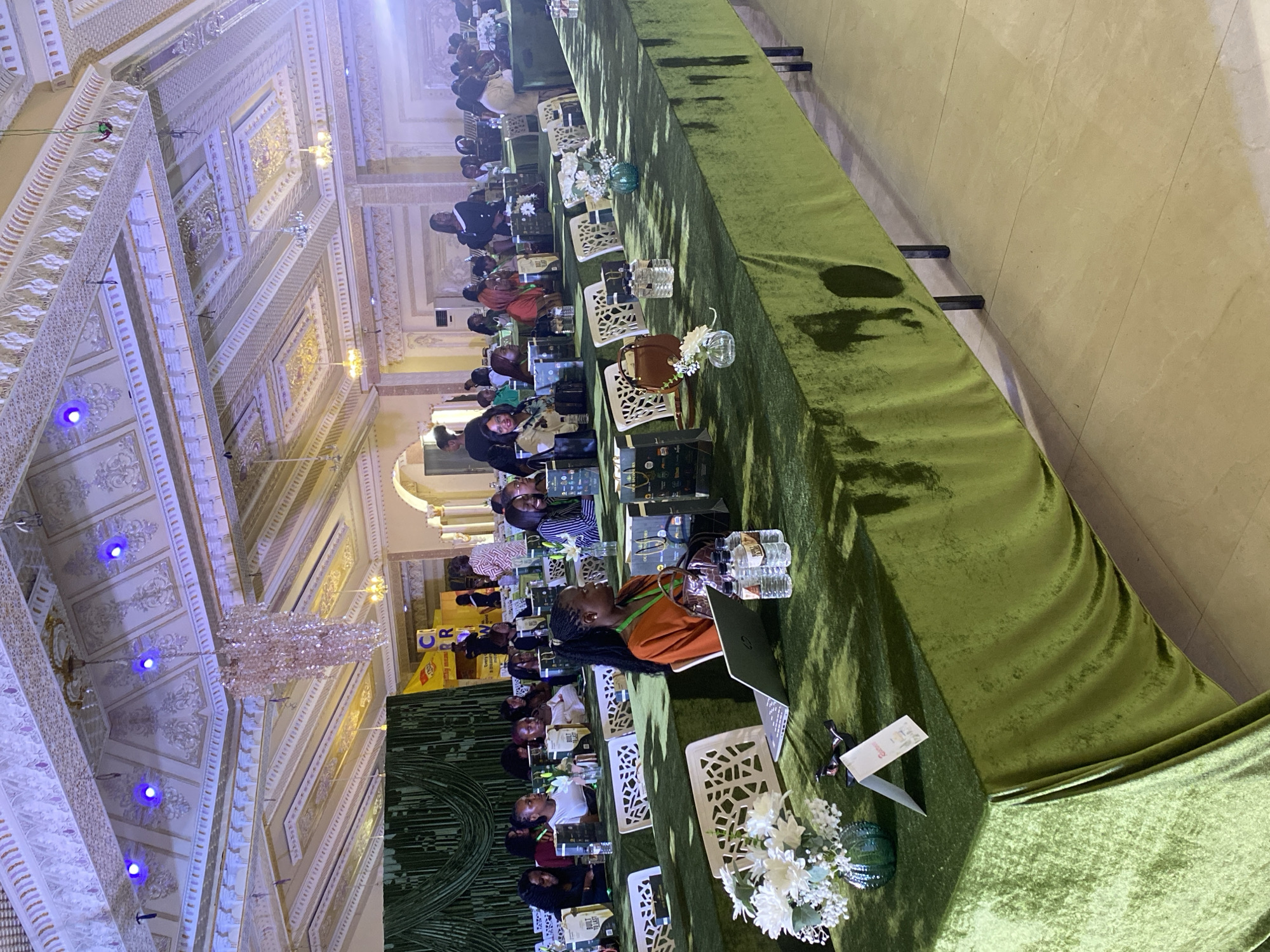 In the city of Jos, Nigeria, an indoor conference or event is taking place within a grand hall. The hall features an elaborately decorated ceiling with intricate moldings, and a large, crystal chandelier hangs from the center. Blue lights are strategically placed within the ceiling's design. Rows of tables draped in green velvet fabric are set up, facing a stage or presentation area that is partially visible on the left side of the image. On the tables, laptops, water bottles, floral arrangements in glass vases, and bags are distributed. A number of women, dressed in various outfits, are seated at the tables, appearing to be attendees at the event. Some are looking forward, possibly at a speaker or presentation, while others are engaged in conversation or reviewing materials. The overall atmosphere suggests a formal gathering or a workshop.