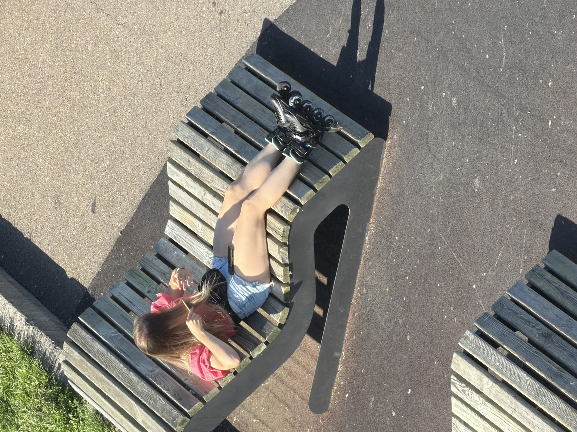 A young female with blonde hair is lying on a wooden lounge chair, wearing a pink shirt, denim shorts, and rollerblades. She is using a comb to brush her hair and appears to be holding a mobile phone in her lap. The scene takes place outdoors on a paved surface, with a patch of grass and concrete in the background. The lighting suggests it is a sunny day. There is no identifiable information suggesting this location is in Krasnoyarsk, Russia.