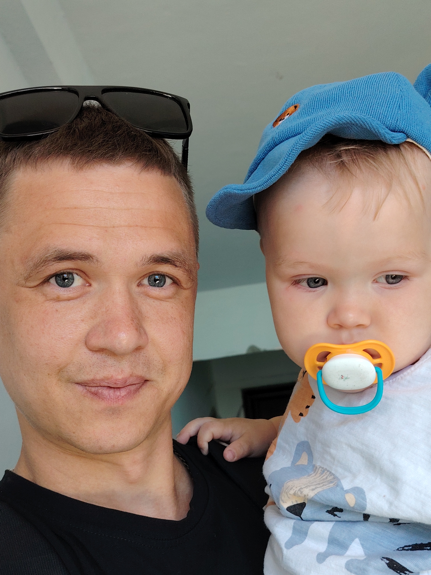 A close-up shot captures a man holding a baby. The man has short brown hair, with sunglasses placed on top of his head, and light-colored eyes. He is wearing a black t-shirt. The baby is wearing a light blue cap with a design on it and a pacifier in its mouth. The baby is wearing a white patterned shirt. The background is a neutral, likely a wall. The location of Chaikovsky, Russia, is unknown.