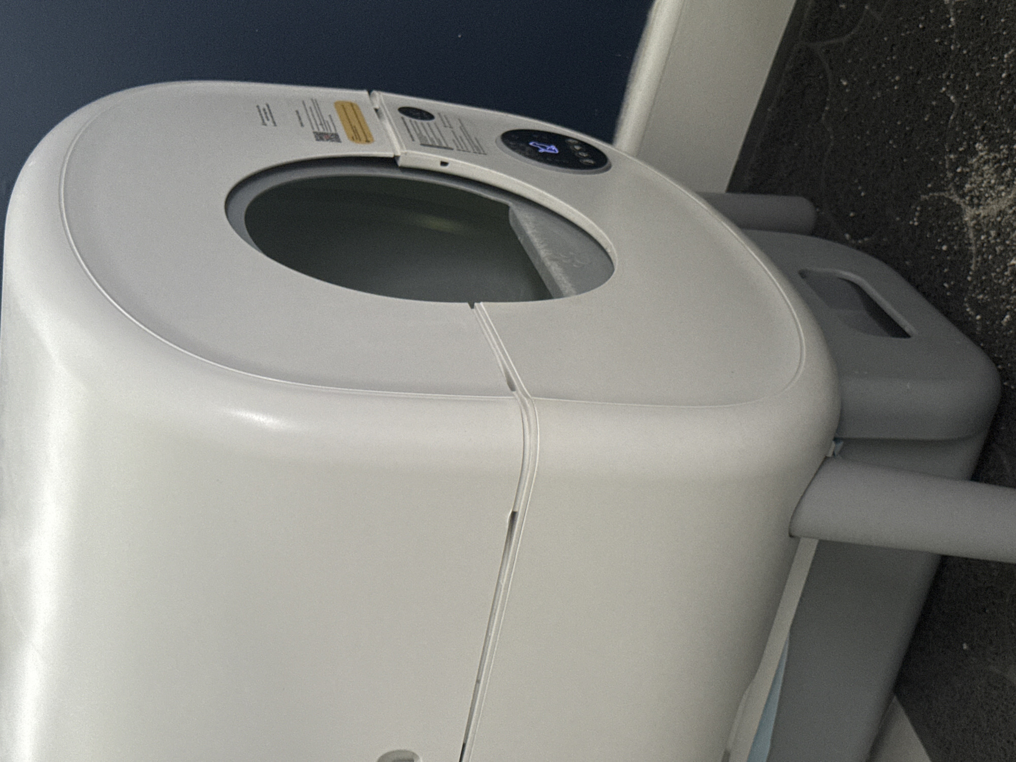 The image displays a white, compact, top-loading appliance, likely a portable washing machine or spin dryer, situated indoors. The appliance is predominantly white with light grey accents and has a sleek, modern design. It features a circular opening on its top surface for loading, suggesting a drum or tub inside.
A control panel is located on the top edge of the appliance, near the opening. This panel includes a small digital display showing "14:00", possibly indicating a timer or program duration. Next to the display, there's a visible lightning bolt icon, typically associated with power or a specific function like spinning. Other unidentifiable buttons or icons are also present. A yellow warning label and another white label with small, unreadable text and a QR code are affixed to the control panel area.
The body of the appliance appears to be constructed from two main sections, joined by a subtle seam running vertically down its side. A light grey handle or support structure is visible on the right side of the unit.
The background behind the appliance is dark grey or black, with a visibly textured surface, sprinkled with numerous small white specks, suggesting dust or debris. There is no visible activity, and the appliance appears to be stationary. The time of day is not discernible, and there are no people present.