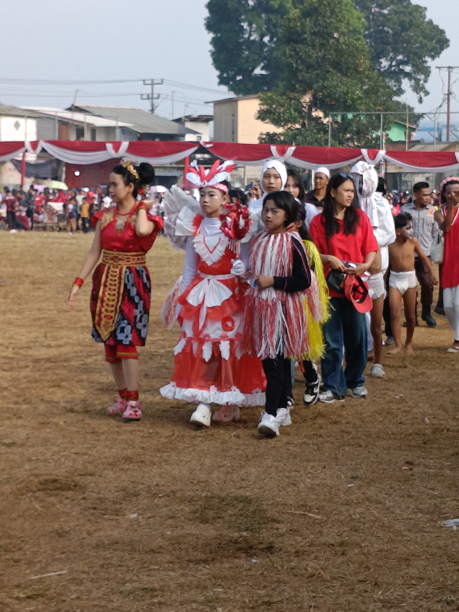 In Cibodas, Indonesia, a group of people are gathered on a dry, grassy field. In the foreground, three young girls in colorful costumes are walking. The girl on the left wears a traditional red and gold outfit with a patterned skirt. The middle girl is dressed in a white and red costume with ruffled layers and a headdress. The girl on the right wears a black outfit adorned with a fringe of red and yellow strands. Behind them, adults and children dressed in everyday clothing and some in uniform are visible. A boy in a white diaper is seen standing near the center. In the background, white and red banners are draped across structures, and trees line the horizon under a hazy sky.