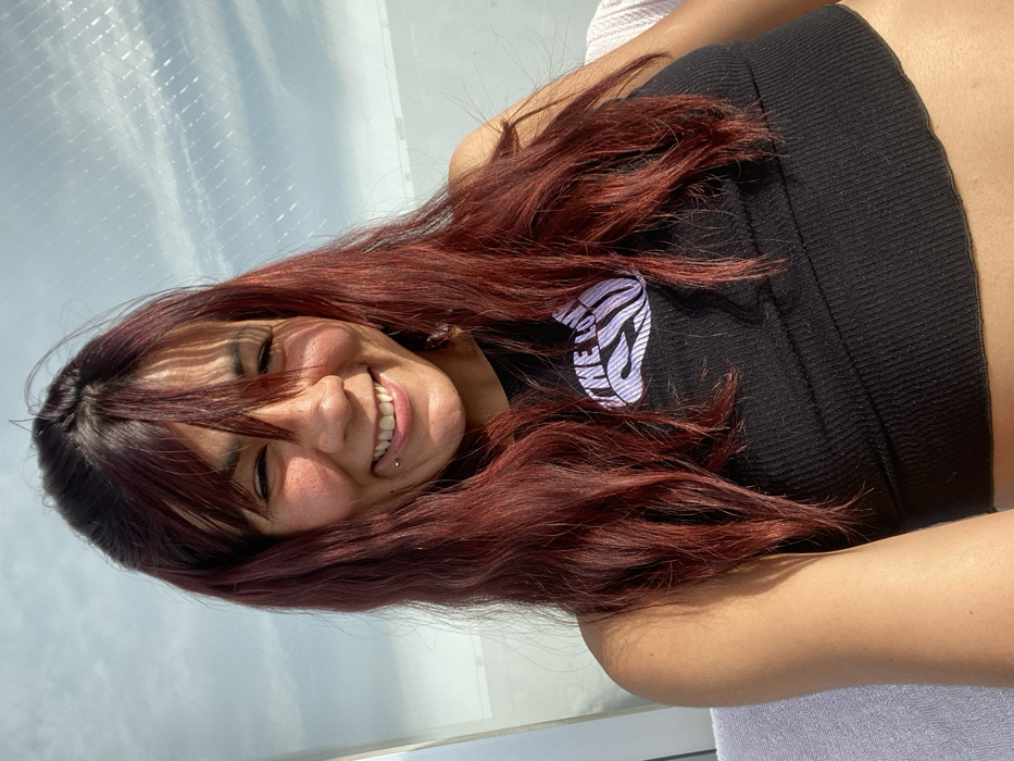 A smiling young woman with red hair and bangs is wearing a black crop top with a white graphic. She has a visible lip piercing. The photo is taken at an angle, with a window or skylight in the background letting in bright light. The light casts shadows across her face and hair. The overall impression is casual and sunny.
