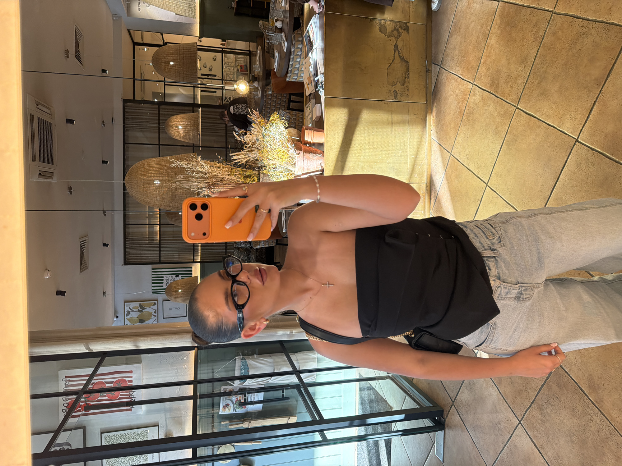 A woman wearing a black strapless top, light blue jeans, and black-rimmed glasses is taking a selfie using an orange phone. She is in front of a glass display case in a restaurant. The restaurant interior has a modern design with wicker lamps hanging from the ceiling, visible in the reflection of the glass. There are decorative elements such as a dried floral arrangement on a table. Artwork is visible in the background, including a tomato-themed painting. The floor is tiled. The restaurant appears to be located in Krasnoyarsk, Russia, based on the writing on one of the paintings.