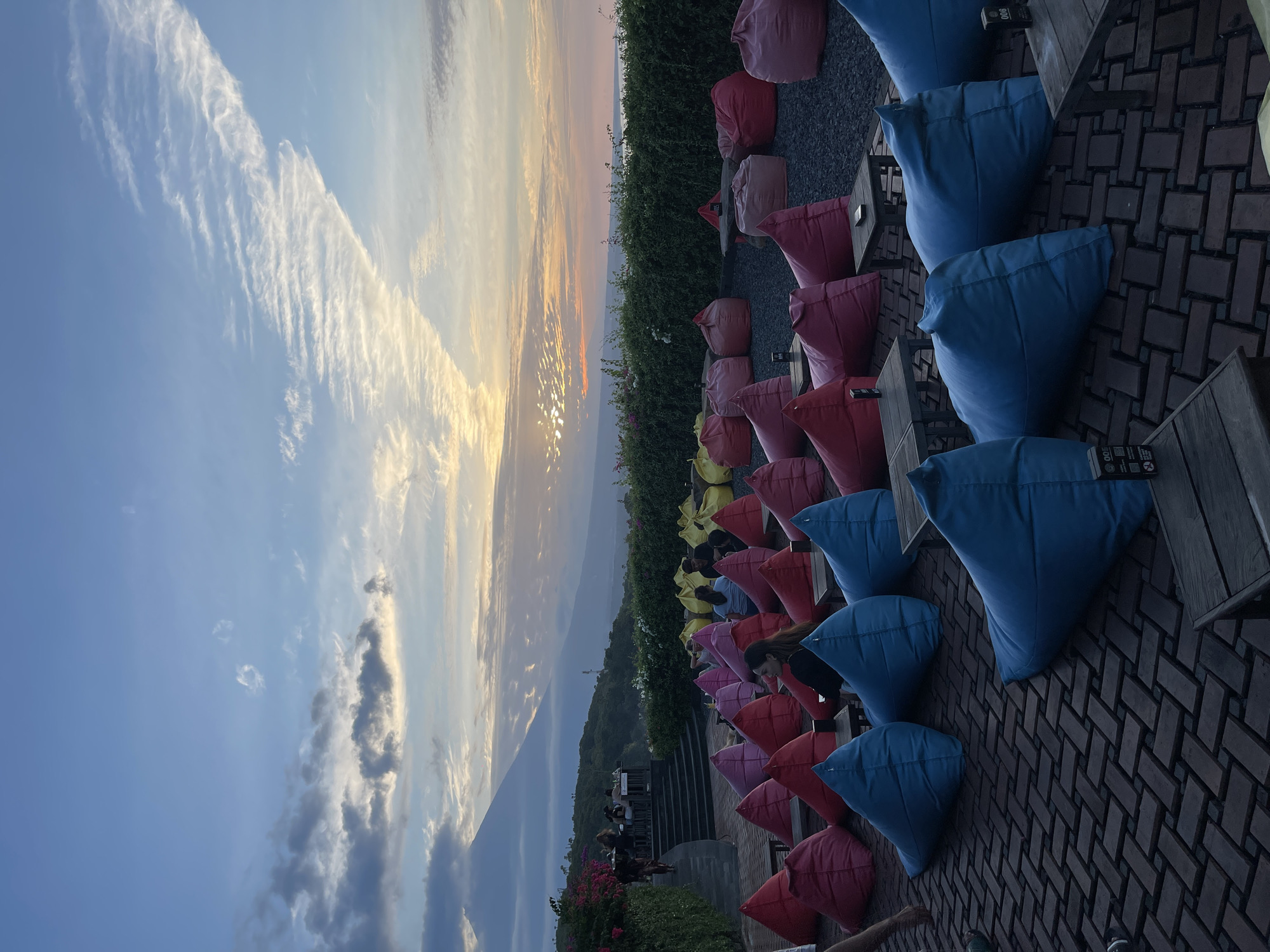 An outdoor seating area with beanbag chairs is set up for a sunset viewing, located in a hilly region overlooking a body of water. In the foreground, numerous large, colorful beanbag chairs in shades of red, pink, blue, and yellow are arranged on a brick patio. Several low wooden tables are interspersed among the beanbags. In the background, a lush green hillside is lined with a dense hedge. Beyond the hedge, a serene ocean view is visible, with mountains in the distance. The sky displays a gradient of soft blue and orange hues, indicating either sunrise or sunset, with wispy clouds scattered across the horizon. A few people are seated at tables on a raised platform to the left, partially obscured by foliage. A single person is walking down a set of stairs on the left side of the image.