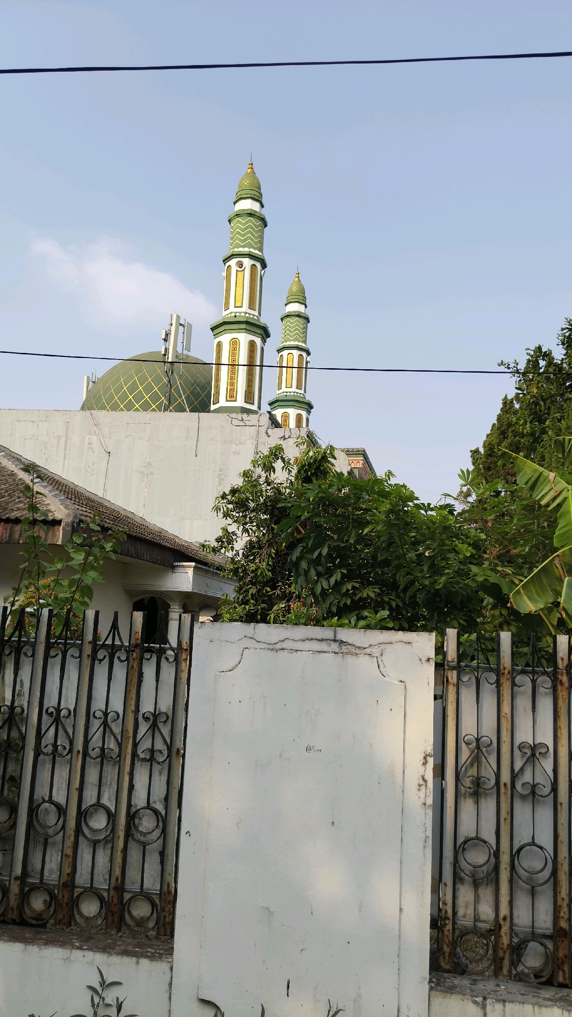 In the city of South Jakarta, Indonesia, a mosque's green dome and two ornate green and gold minarets are visible above a wall and a rusty metal fence. The minarets have decorative bands and pointed finials. The mosque is situated behind a residential building with a grey roof. Lush green trees and foliage obscure parts of the lower structure of the mosque and the surrounding area. Overhead, black electrical wires are strung across the frame against a pale blue sky with some scattered clouds. The foreground consists of a white wall and a section of the fence, indicating a street-level view.