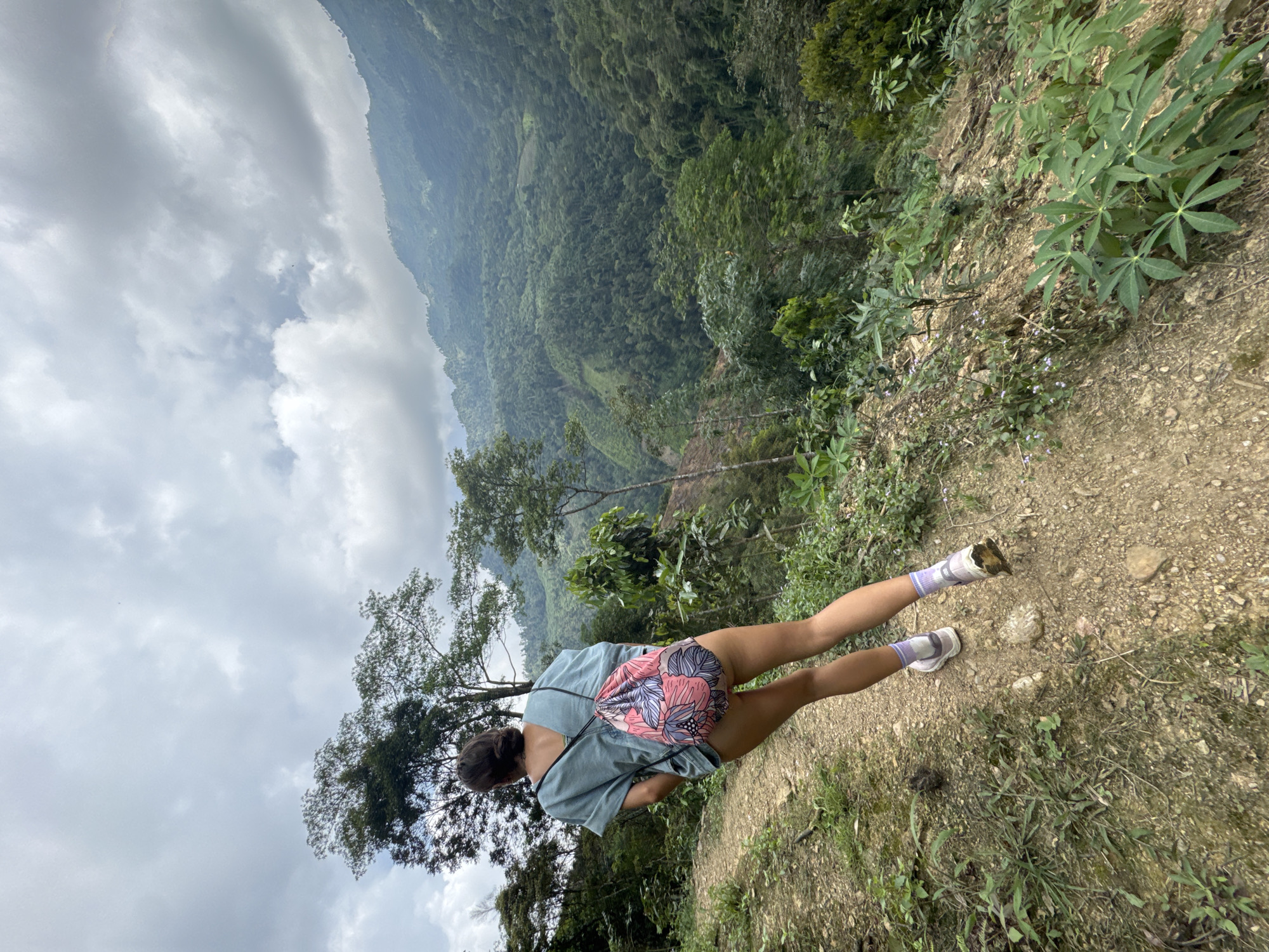 A woman stands on a dirt path in Suối Giàng, Vietnam, facing a forested valley under a cloudy sky. She wears a light blue shirt, shorts, lavender socks, and sneakers. A floral backpack is slung over her shoulder. The path is flanked by vegetation, and a steep drop-off is visible. The landscape is lush and green, and the sky is overcast.