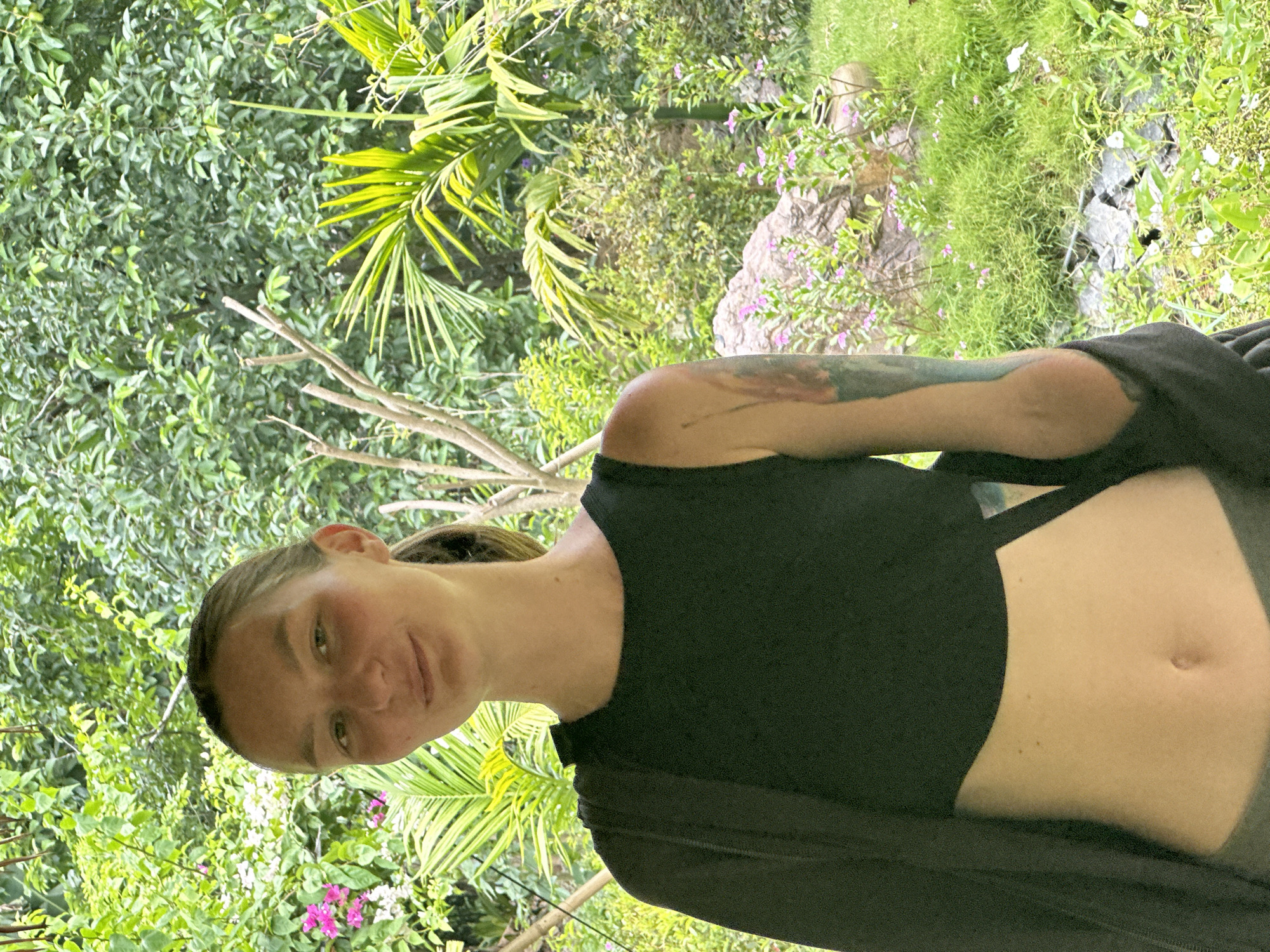 A young woman with blonde hair pulled back in a ponytail and a faint smile stands in front of lush greenery. She is wearing a black crop top and a dark overlay garment draped over her left shoulder. Her left arm is visible, displaying a tattoo. The background is filled with dense foliage, including palm fronds and pink flowers, suggesting a tropical or garden setting. A large rock and scattered stones are visible to her right, interspersed with grass and more vegetation. The lighting suggests it is daytime, likely outdoors. The woman appears to be posing for the camera. The scene is located in the city of Nam Hoa Lư Ward, Vietnam.