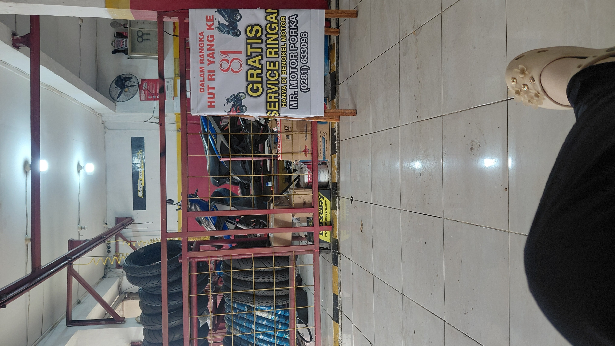 The image depicts the interior of a motor vehicle workshop, possibly in Bantarsoka, Indonesia, with a focus on a promotional banner. The banner, displayed on a wooden easel, advertises "GRATIS SERVICE RINGAN" (Free Light Service) in celebration of Indonesia's 81st Independence Day ("HUT RI YANG KE 81"). It also lists the workshop's name as "MR. MOTOR PORKA" and provides a contact phone number, (0281) 633096.
Behind the banner, two motorcycles are parked. The workshop area is organized with racks of tires visible on the left. Various boxes and containers are also present, some appearing to be for oil or spare parts. The floor is tiled in a light color. In the foreground, a person's legs and feet are visible, suggesting they are seated and the photo is taken from their perspective. One foot is wearing a light-colored shoe adorned with floral embellishments. The environment appears to be a functional, working garage space.