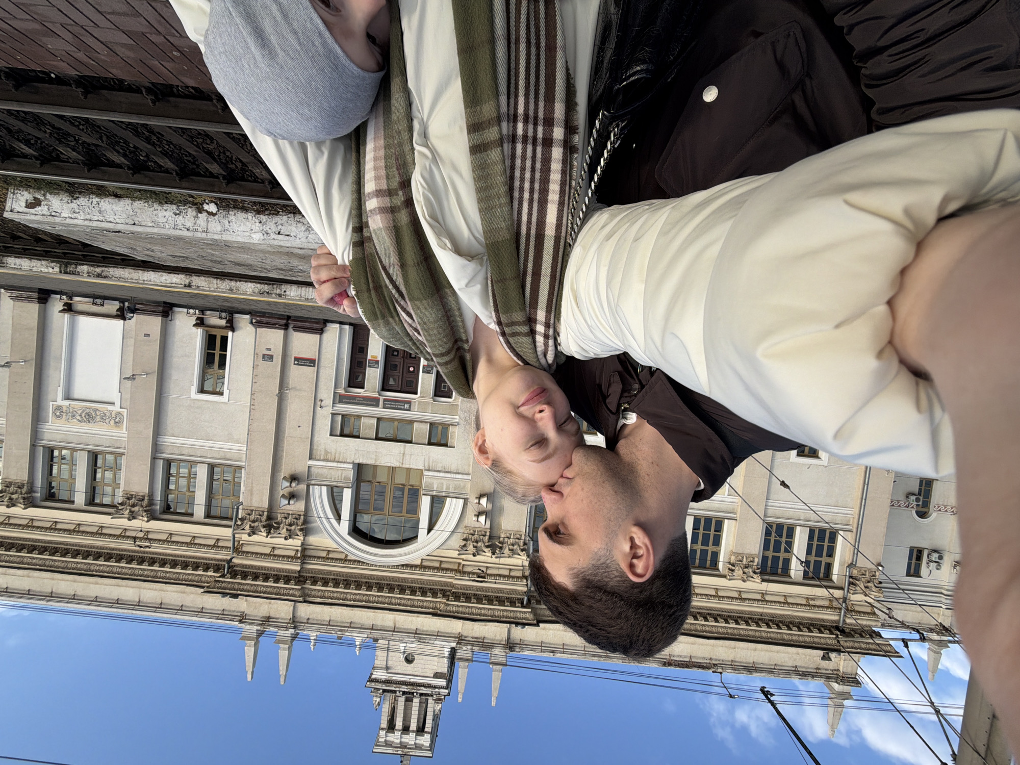 A man and a woman are shown in a close-up selfie. The man is kissing the woman on the cheek. The woman has light skin, and she is wearing a gray hat, a beige jacket, and a plaid scarf. The man has dark hair and is wearing a dark jacket.
Behind the couple is a building with a clock tower. The building appears to be the Krasnodar-1 Train Station, located in Krasnodar, Russia. Above the building is a blue sky with some clouds. Wires are visible in the foreground.