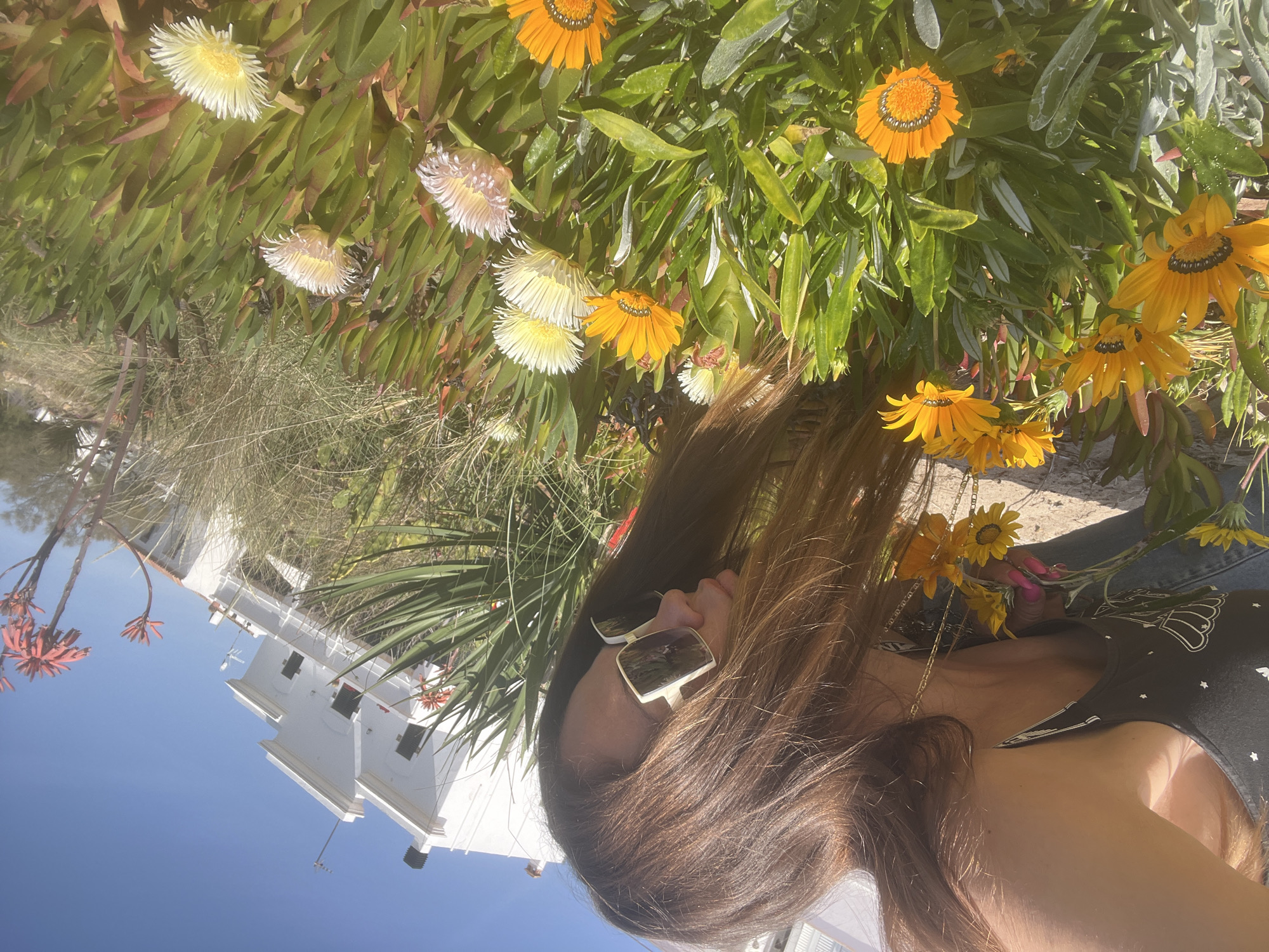 A woman in sunglasses stands in front of a mix of yellow and white flowers, with a white building in the background under a clear blue sky. The location is likely in La Marina De Creixell, Spain.