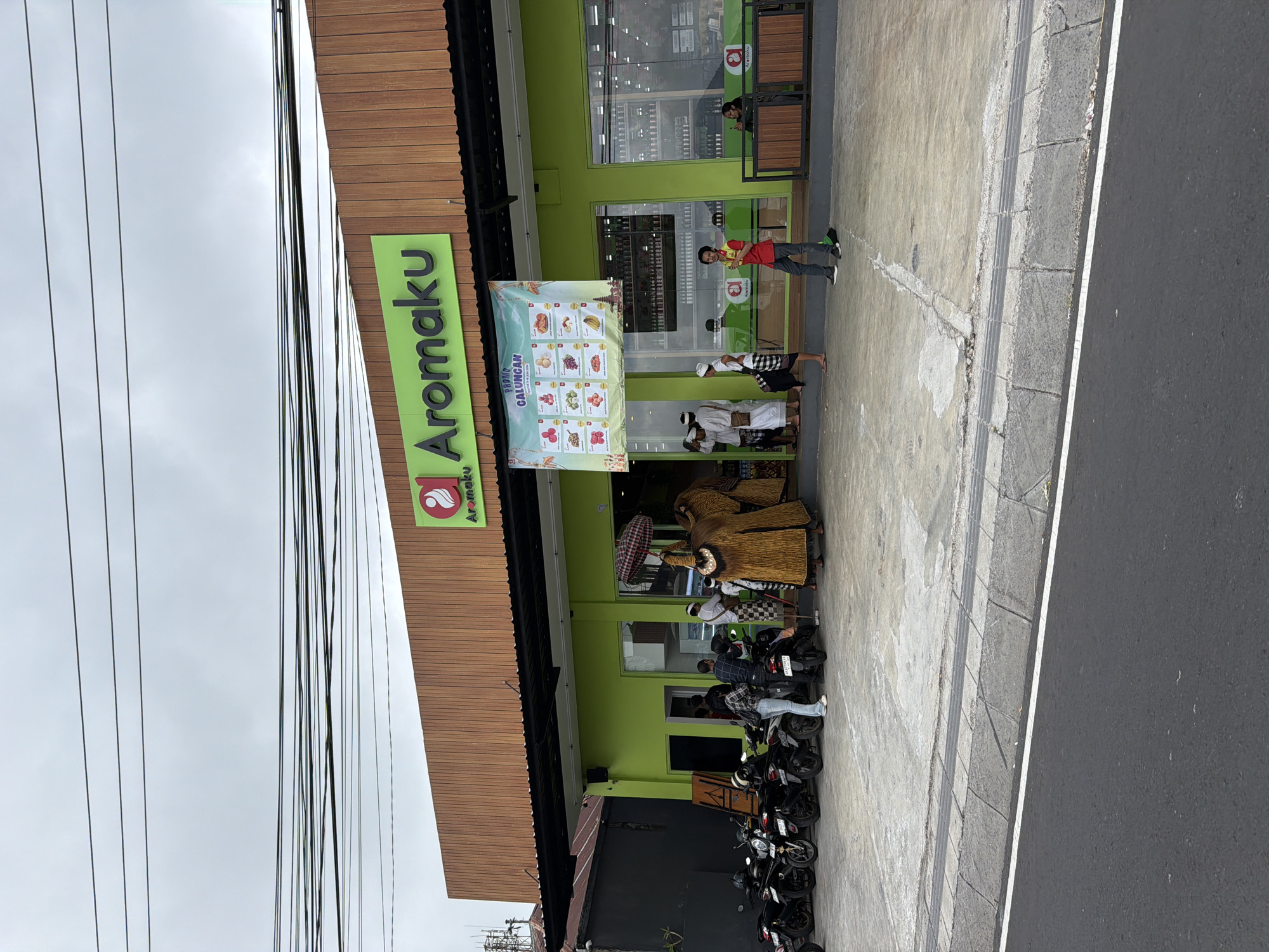 The image depicts the front exterior of a bright green store named "Aromaku" in Kintamani, Indonesia. The store has a wooden-paneled facade above the green storefront. A banner promoting "PROMO GALUNGAN" hangs across the entrance, showcasing various food items. Several people are present outside the store. Some individuals in traditional Balinese attire, possibly part of a cultural event, stand near the entrance. Others are seen near a row of parked motorcycles to the left of the store. A man in a red shirt and dark pants is standing near the entrance, and another person is seated on a bench to the right. The scene suggests a bustling environment with both shopping and cultural activities. The sky is overcast, and the ground is paved.
