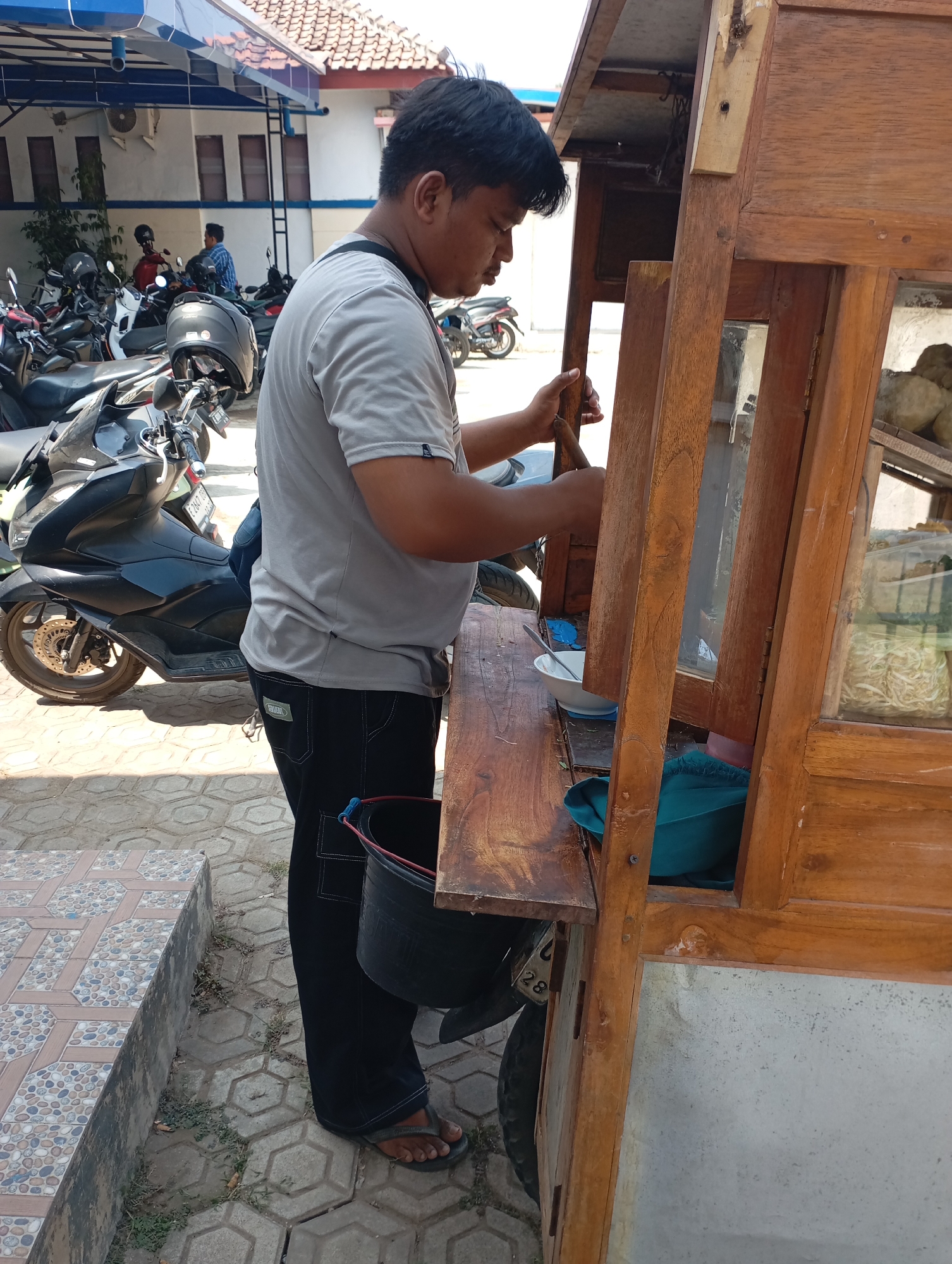 An Indonesian man, dressed in a grey t-shirt and black pants, stands beside a wooden food cart. He is holding a wooden utensil in his right hand and appears to be serving food from a bowl. The food cart has a glass display case showing ingredients. In the background, several motorcycles are parked, and a building with white walls and a tiled roof is visible. The ground is paved with interlocking concrete blocks. The scene suggests an outdoor food stall in a bustling area of Kecamatan Haurgeulis, Indonesia.