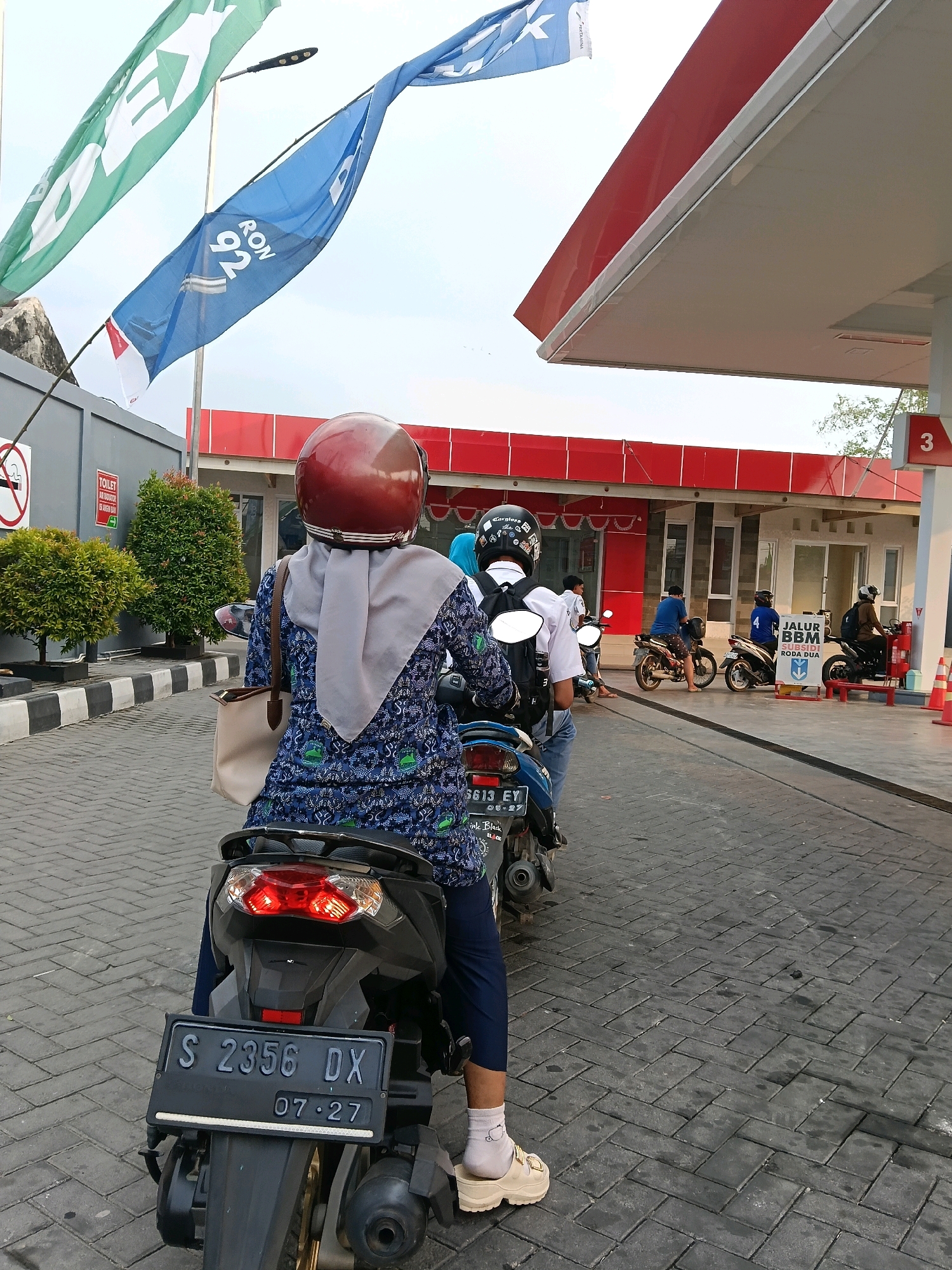 In Tambakromo, Indonesia, a person wearing a red helmet and a blue and green patterned dress is seated on a motorcycle, viewed from behind. The motorcycle is positioned in a queue at a Pertamina gas station. The scene captures a sunny day with flags flying, including a blue flag with "RON 92" and a green flag with a white arrow. The gas station features red and white architecture, and a sign in Indonesian indicates "JALUR BBM KHUSUS RODA DUA" (Fuel Lane for Two-Wheeled Vehicles). Other individuals on motorcycles are also visible in the queue, waiting to refuel. The ground is paved with interlocking grey bricks.