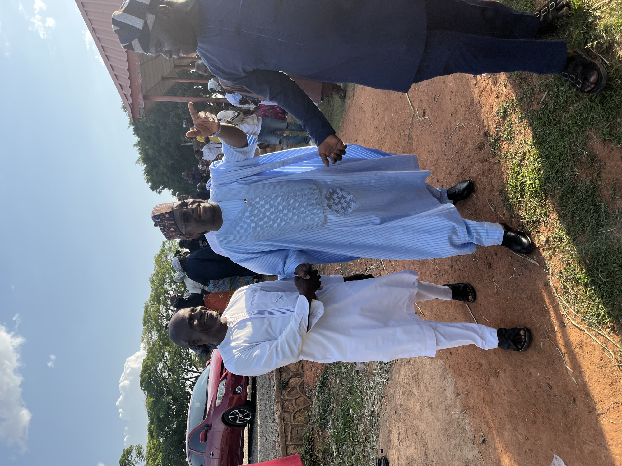 The image captures a scene in Jos, Nigeria, showing three men outdoors on a sunny day, likely at an event. The man in the foreground wears a white traditional outfit and black sandals, standing beside a red car parked along a low stone retaining wall. Behind him, a man in a light blue striped traditional robe and black shoes gestures with his right hand, seemingly addressing a crowd visible in the background. Partially obscuring the view is a third man in a dark blue outfit and sandals, standing closest to the camera. The ground is a mix of red soil and patchy grass. A building structure with a red roof is visible in the background, further indicating an outdoor gathering. A partly cloudy blue sky is above.