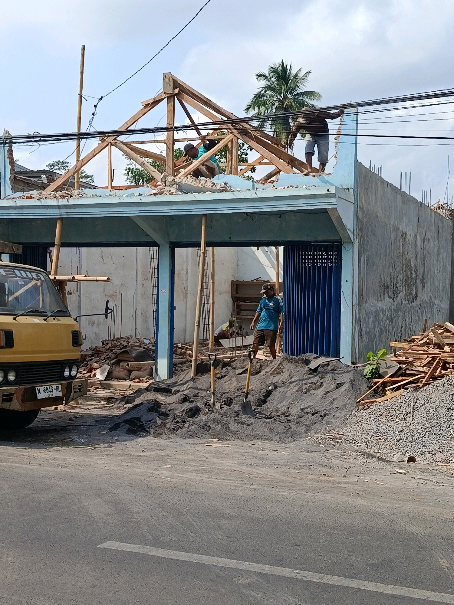 In Sawaran Lor, Indonesia, a construction site is actively undergoing demolition and rebuilding. The image captures an overcast sky above a partially constructed or deconstructed building. A yellow truck is parked to the left, its license plate N 8843 RL visible. The building itself features a light blue overhang and concrete walls. The roof structure is made of exposed wooden beams, with two men working on top. One man, wearing a blue shirt and dark shorts, is crouched and actively engaged with the wooden framework. Another man, in a dark shirt and light shorts, stands further back on the roof, also appearing to be working. Below, a pile of dark sand and gravel dominates the foreground, with a shovel embedded in it. Debris, including bricks and pipes, is scattered around the base of the building. A closed blue metal gate is visible on the ground floor. In the background, a palm tree stands tall against the sky, and electrical wires crisscross overhead. The scene suggests ongoing construction work with workers engaged in manual labor.