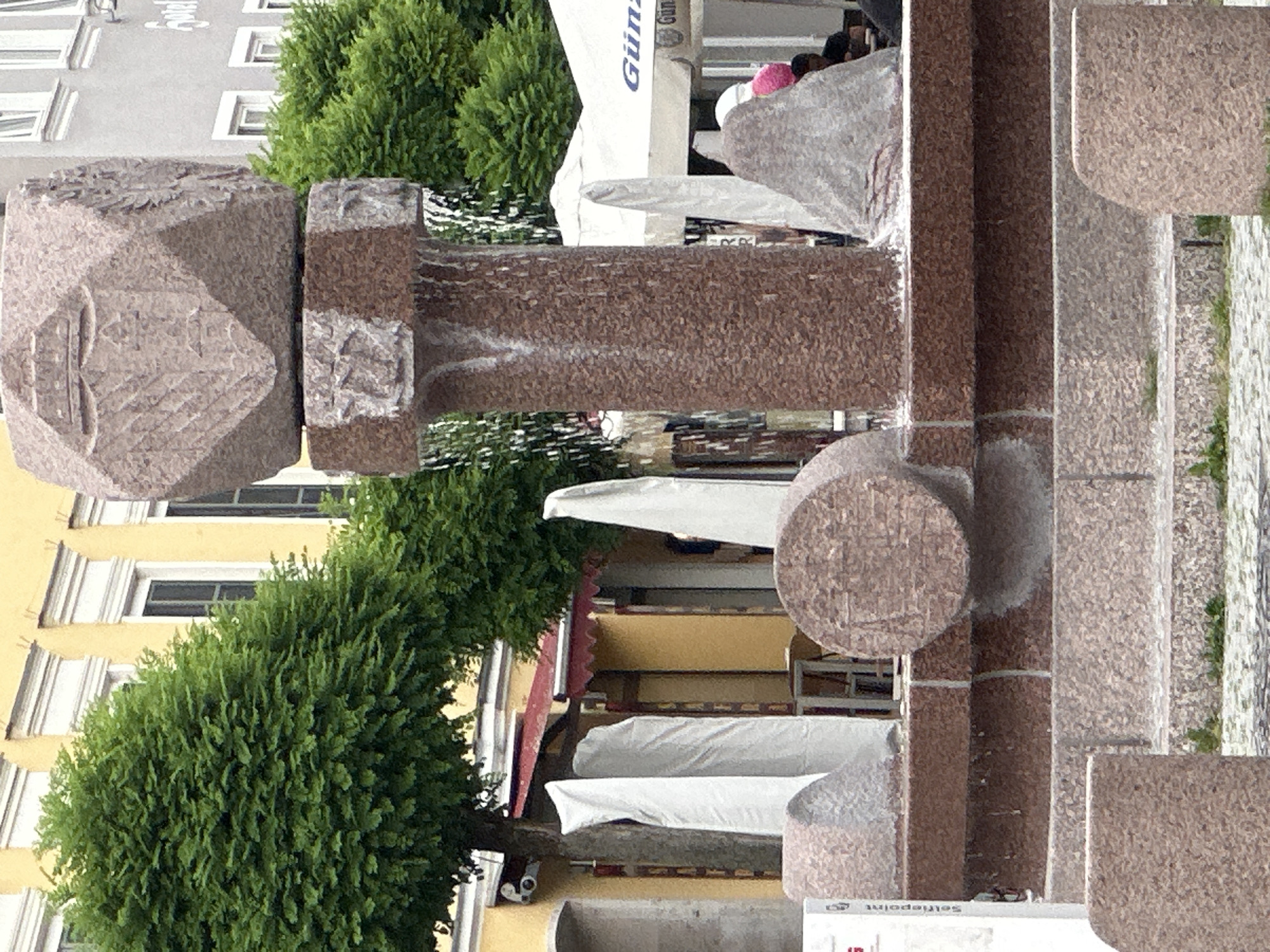 A stone fountain is the central focus of this outdoor image, set in a public space in the city of Unknown. The fountain is constructed from pinkish-brown granite and features a complex design with carved details and water flowing down its sides.
The fountain's structure includes several stacked geometric shapes, with the uppermost element resembling a faceted stone bearing a carved emblem or crest. Below this is a cylindrical column with water cascading from its top. The base of the fountain is a tiered structure with a circular basin, and various other stone elements, some covered in fabric, are situated around it.
Behind the fountain, a yellow building with white-trimmed windows is visible, suggesting an urban environment. Lush green trees with dense foliage frame the scene, and a red awning is partially visible on the yellow building. In the background, white umbrellas with branding like "Günz" indicate an establishment, possibly a cafe or restaurant. A few people are present in the background, one wearing a pink hat, and others seated under the umbrellas. The ground around the fountain is paved with cobblestones and some patches of grass. The overall impression is that of a public square or plaza.