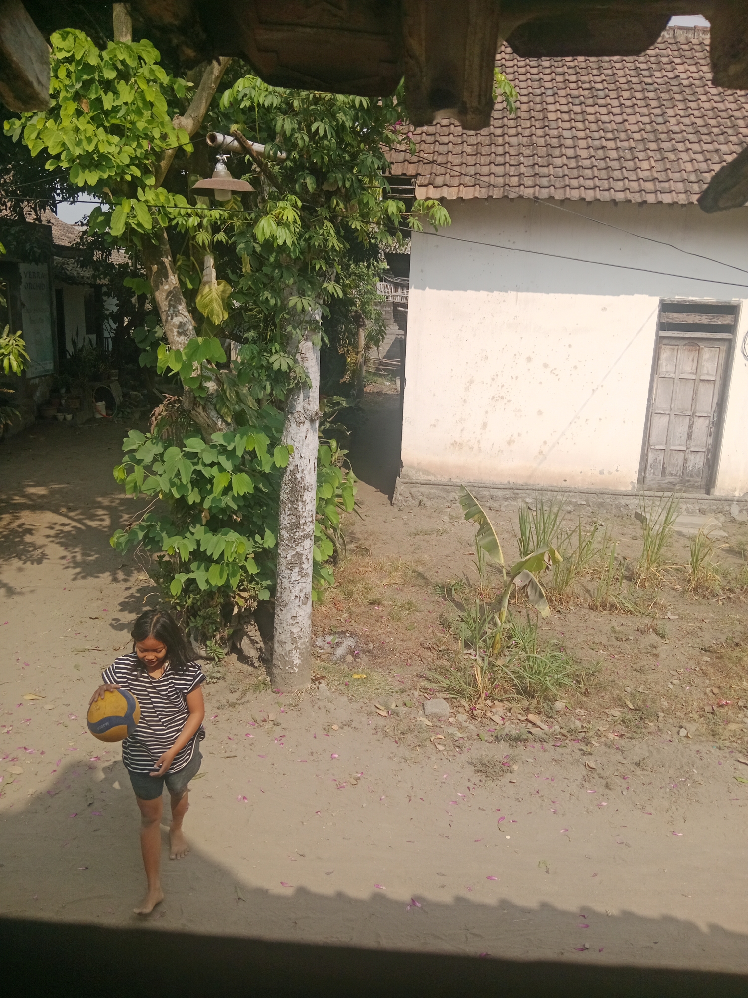 A young Indonesian girl stands in a dirt yard in front of a modest house in Ringinrejo, Indonesia. She is holding a volleyball and is wearing a striped t-shirt and shorts. The house has a tiled roof and a wooden door. There are trees and vegetation around the yard. The image appears to have been taken from an elevated position, possibly a window or balcony, looking down at the scene.