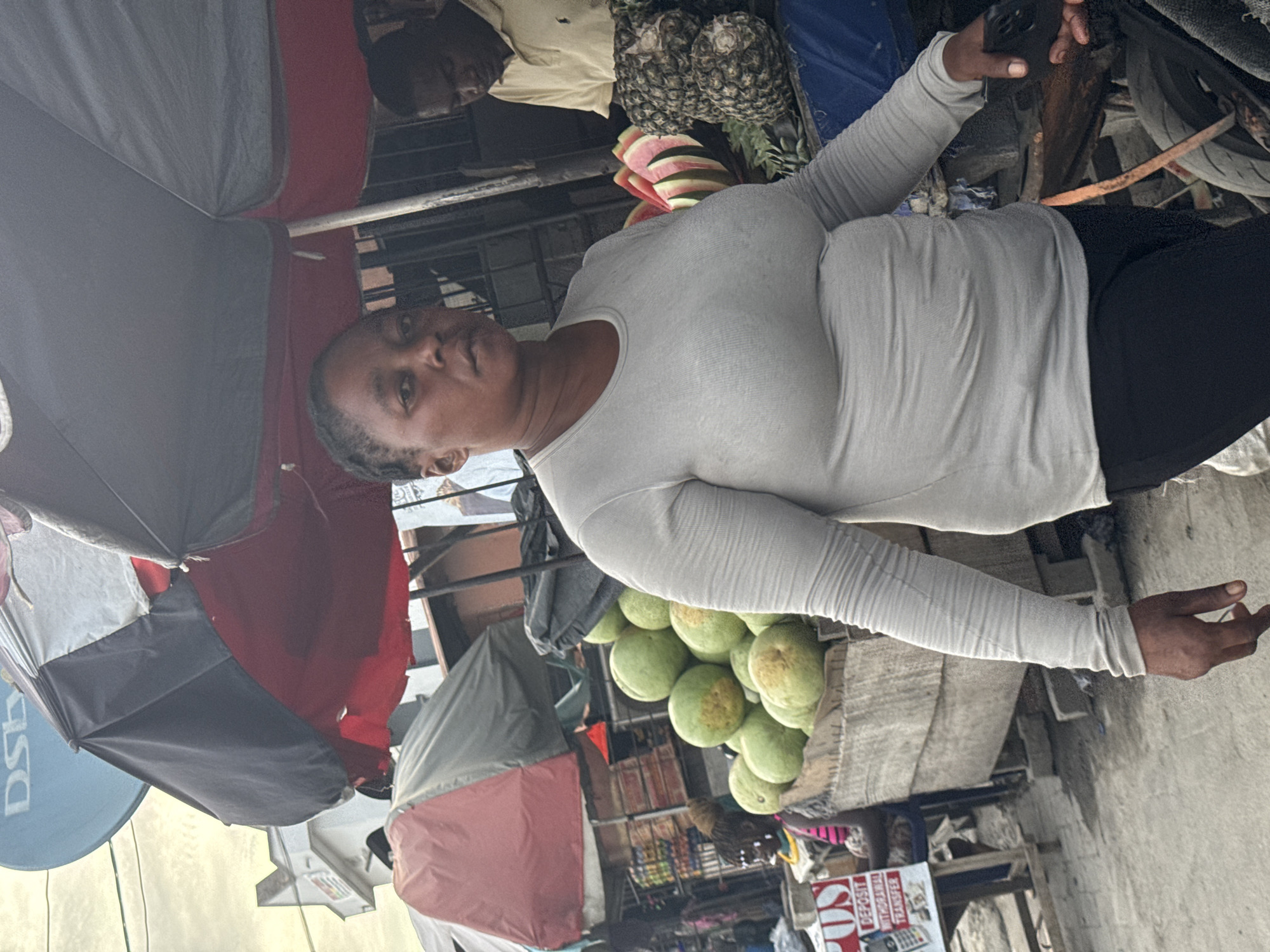 The image depicts a woman in a light-colored long-sleeved shirt and dark pants standing behind a fruit stand. The fruit stand contains a pile of round green fruits, possibly melons. Behind her, there are pineapples and cut watermelon. She is holding a dark object, possibly a mobile phone, in her right hand. The scene appears to be an outdoor market or street-side vending location. Several umbrellas are visible, some red and white, possibly shading stalls. Another man in a light-colored shirt is visible behind the fruit stand. A blue satellite dish is visible in the background, and signs indicate financial transaction services. Based on the general appearance and architectural style, the location is likely in a densely populated area in Lekki, Nigeria.