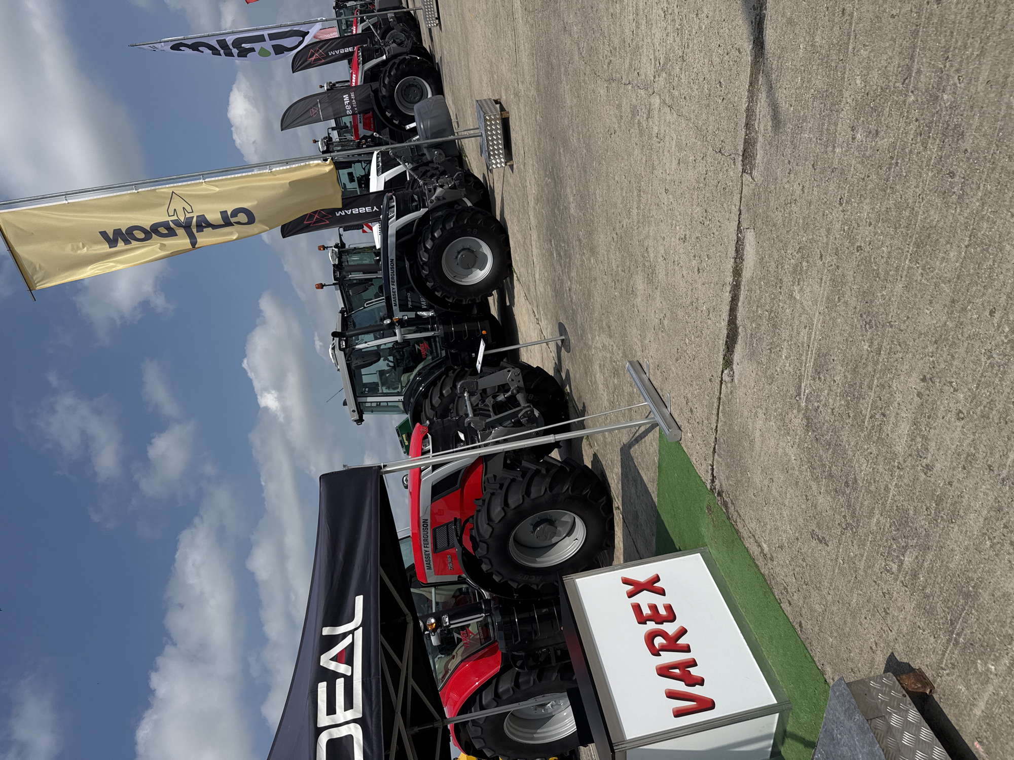 The image captures an outdoor agricultural machinery exhibition, likely in Stara Zagora, Bulgaria, under a bright, partly cloudy sky. Several large tractors are displayed in a row on a concrete hardstanding area.
In the foreground, a red and black Massey Ferguson tractor, with "7S155" visible on its side, is parked next to a black tent or display banner bearing the word "DEAL" in white. Adjacent to this, a white sign prominently displays "VAREX" in red letters, placed on a strip of artificial green grass that borders the concrete pavement.
Further down the line, another Massey Ferguson tractor, primarily grey and black with the model number "7726S" visible, is also on display. Above the tractors, several banners and flags are visible against the blue sky. A prominent yellow banner features the name "CRAYDON" and an arrow logo. Other banners include "MASSEY FERGUSON" and additional brand names such as "BIM," "MAREK," and "MECHEL."
The setting is an open-air exhibition space, illuminated by bright daylight, suggesting a mid-day event. The sky shows scattered white clouds, indicating fair weather. No individuals are visible in the scene. The arrangement of various tractors and multiple company banners strongly points to a trade show or promotional event for agricultural equipment.