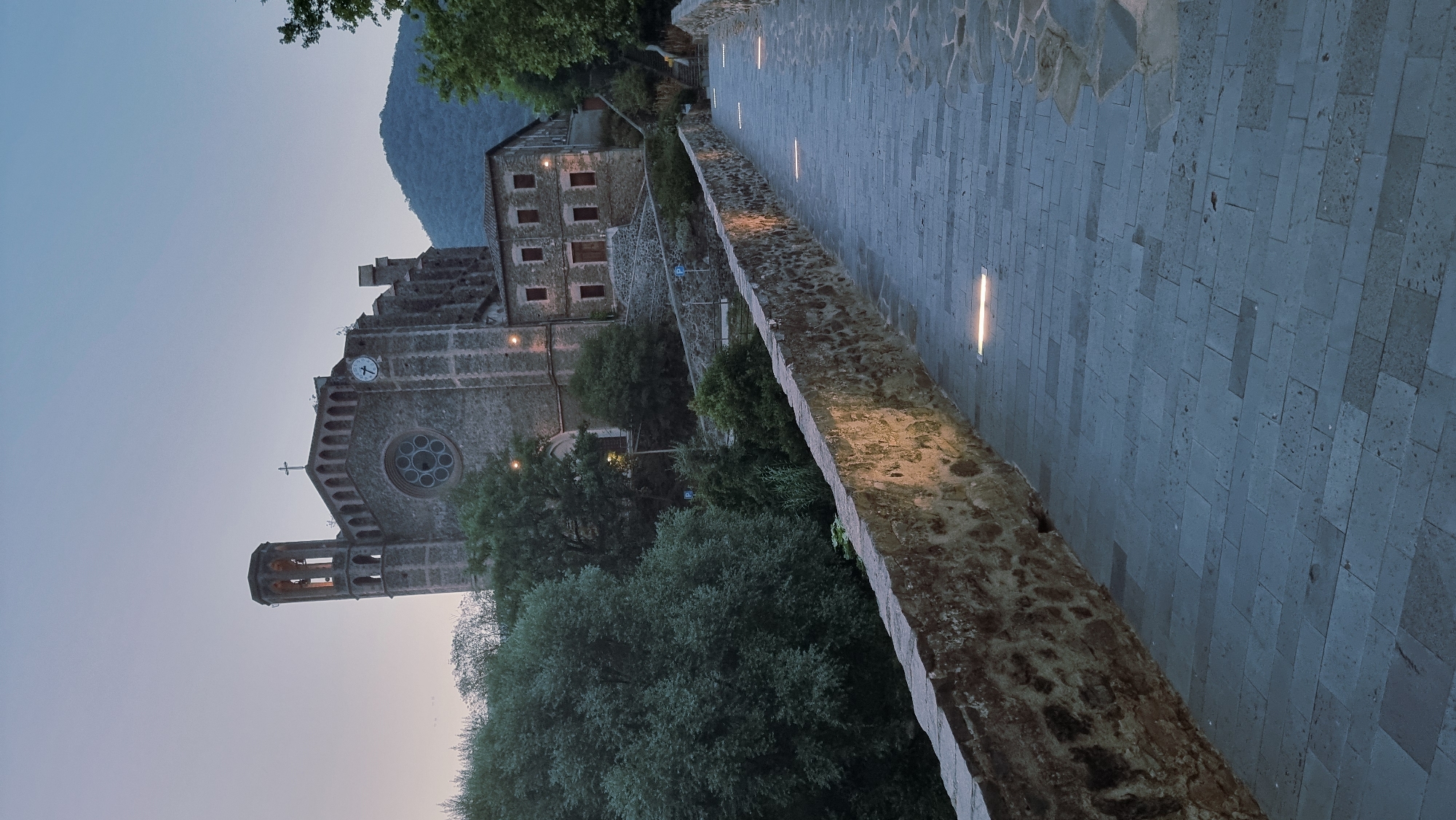 The image shows a stone bridge with embedded lights leading towards a large stone building, possibly a church, in the city of Sant Joan Les Fonts, Spain. The building has a clock on its facade and a tower. The sky is dusky, indicating it is either dawn or dusk. Lush green trees are visible on the left side of the image and behind the building. The bridge has a stone wall on the left, illuminated by the embedded lights that create a warm glow on the stone surface. The pathway of the bridge is paved with large, rectangular stone blocks. The scene depicts an architectural and natural landscape under subdued lighting conditions. There are no people visible in the image.