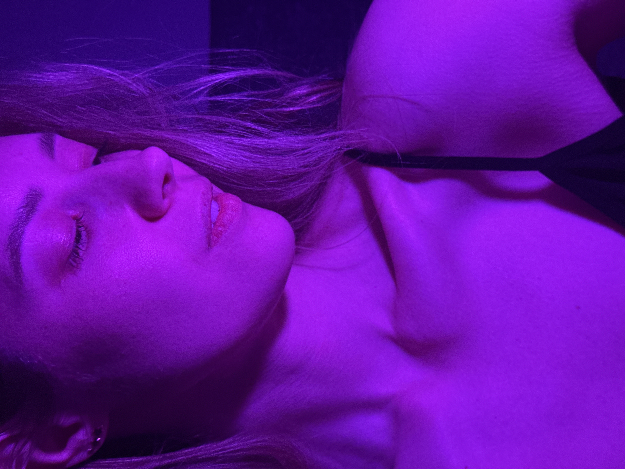 A female individual is lying down, illuminated by a purple light. Her face is turned towards the camera. She is wearing a black top with thin straps. Her hair is light colored. The background is dark. It's not possible to ascertain the location as Chaikovsky, Russia based on visual data alone.
