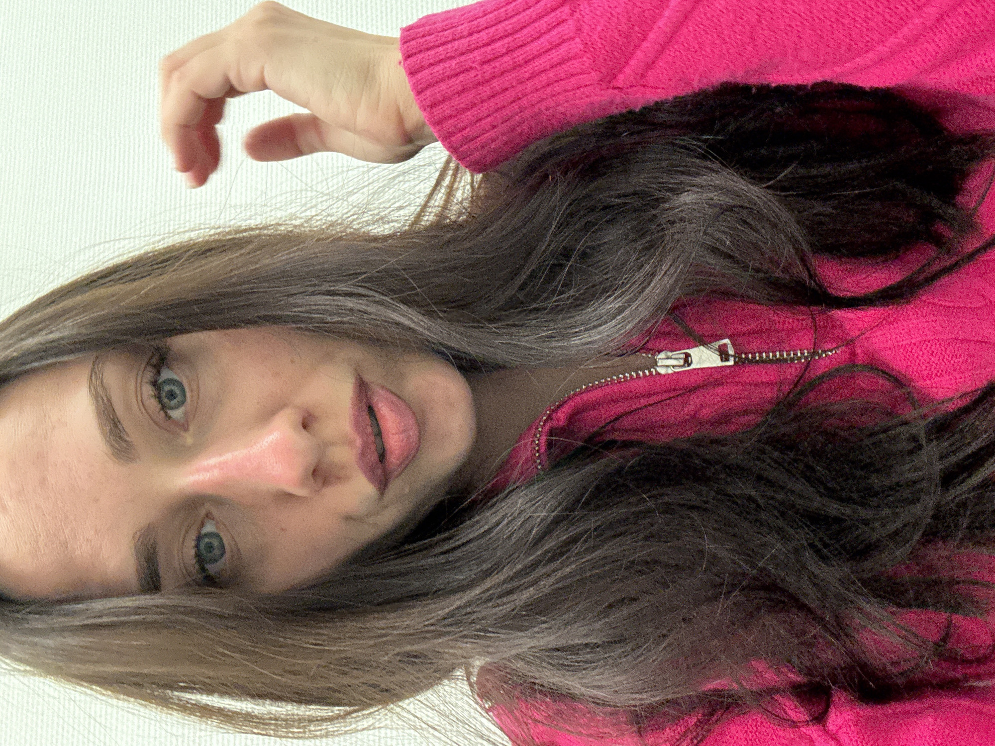 A close-up, rotated portrait captures a person with long, wavy dark brown hair and clear blue eyes, looking directly at the viewer. The image is oriented sideways, featuring the person's head tilted towards the left of the frame. They are wearing a vibrant pink, ribbed sweater, possibly knitted or fleece, with a silver quarter-zip visible on the right side.
The person's lips are slightly parted, revealing a touch of pink lip color, and their expression is neutral to slightly engaged. Their skin is fair. In the upper left portion of the image, one of their hands is visible, with neatly trimmed nails, curved as if resting or gesturing. The background is a plain, light-colored, subtly textured surface, suggesting an indoor setting. No specific time of day or weather is discernible from the image. No text is visible. The location context is Saint Petersburg, Russia.