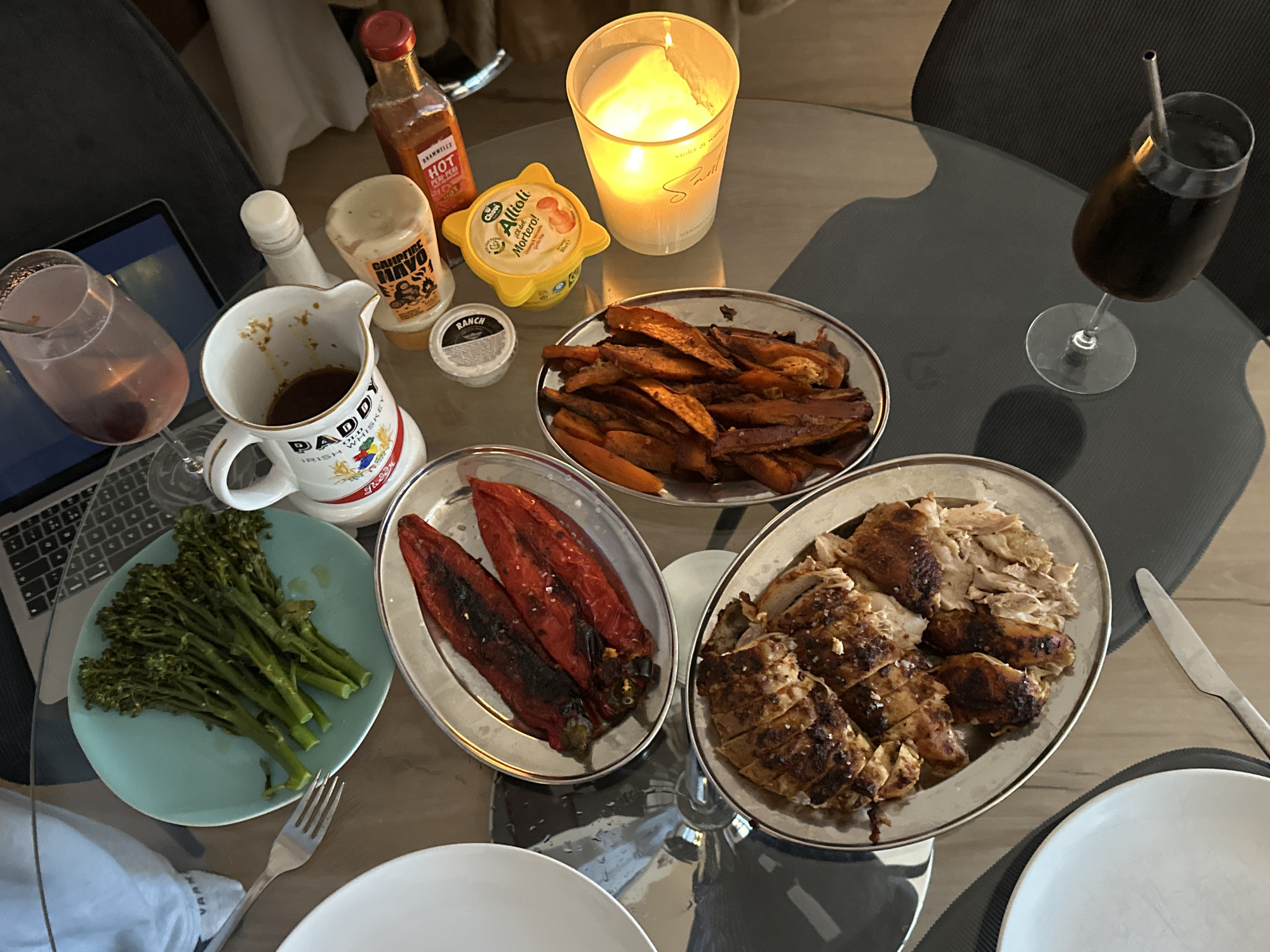 This image depicts a table setting with a variety of food items, drinks, and other objects. The setting appears to be indoors, possibly in London, United Kingdom, considering the presence of "Paddy Irish Whiskey".
On the table, there are several dishes:
* A plate with charred red peppers.
* A plate of roasted sweet potatoes.
* A plate of grilled chicken.
* A plate of broccoli.
Other items on the table include:
* Condiments such as hot sauce, aioli, ranch dressing, and a bottle of "CAUPFIRE MAVO".
* A mug labeled "PADDY IRISH WHISKEY" filled with liquid.
* Two beverages in glasses, one of which has a metal straw.
* A lit candle.
* A laptop computer.
* A knife and fork.
* Two white plates.
The table itself is made of glass. The image presents a scene suggestive of a meal in progress, possibly at home.