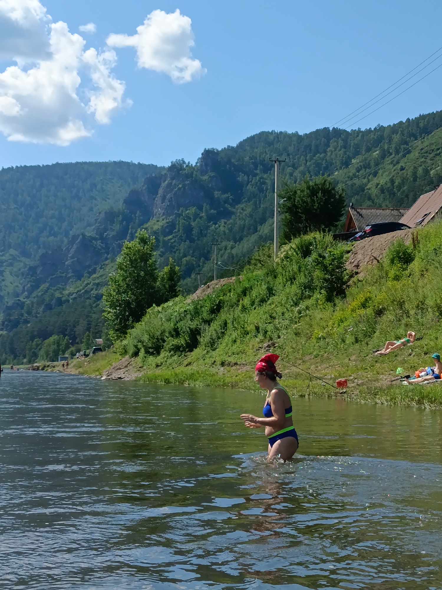 In Усть-Мана, Russia, a young woman wearing a blue and green bikini and a red bandana is standing in a river, with her legs submerged in the water. She is facing to the right, with her hands raised and her mouth slightly open. The river is calm with ripples on the surface. In the background, a grassy embankment slopes upwards, dotted with green bushes and trees. Several people are visible on the embankment; two are lying down, appearing to sunbathe, while others are standing. A few small structures, possibly houses, are situated on the higher parts of the embankment. A power line runs diagonally across the scene, supported by utility poles. In the distance, a large, green, forested mountain range rises under a bright blue sky with scattered white clouds.