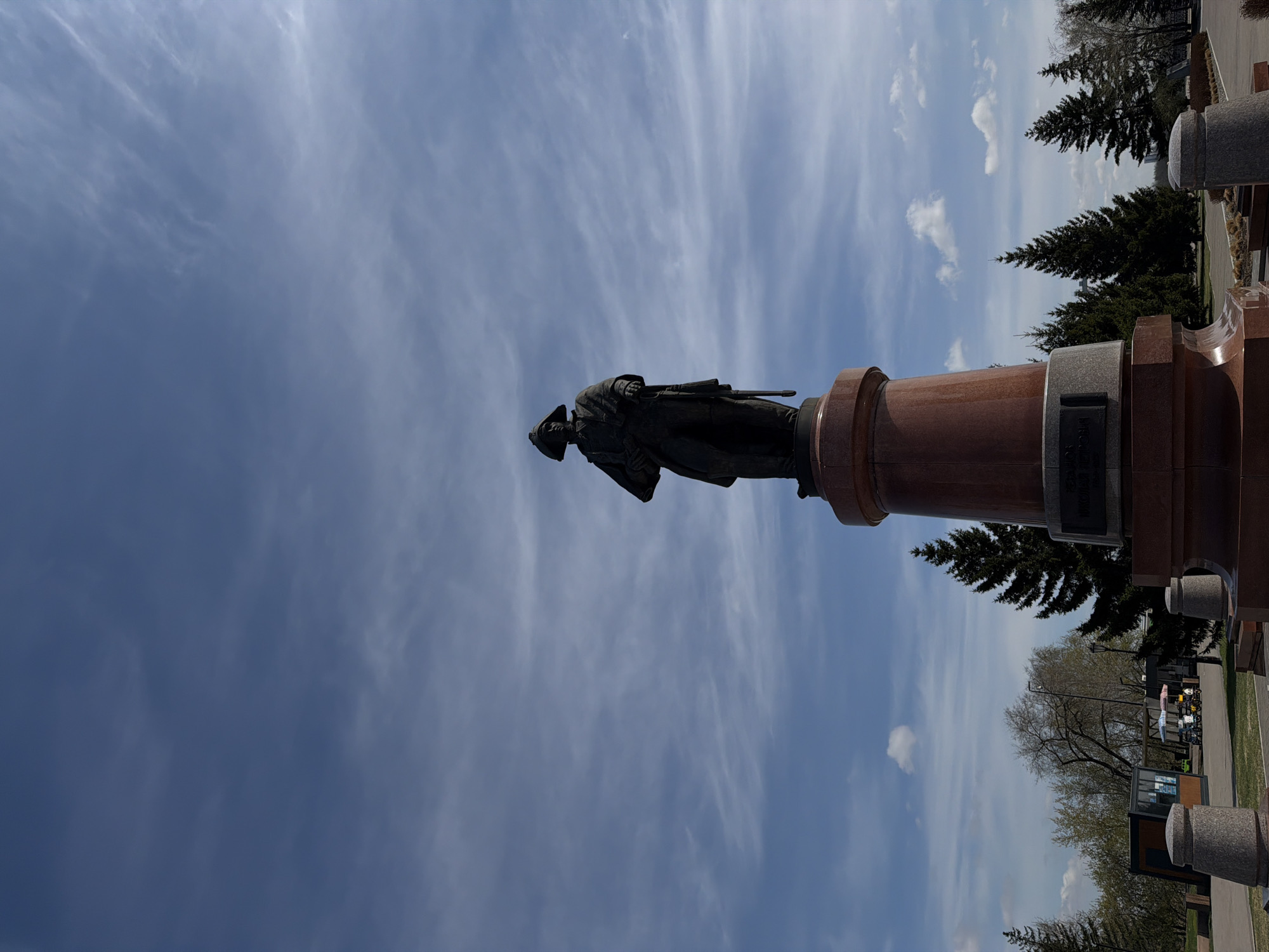 The image shows a statue of a soldier with a rifle on a tall red granite base, located in Krasnoyarsk, Russia. The statue stands in a park or cemetery setting, with trees and a walkway visible. The sky is blue with scattered clouds.