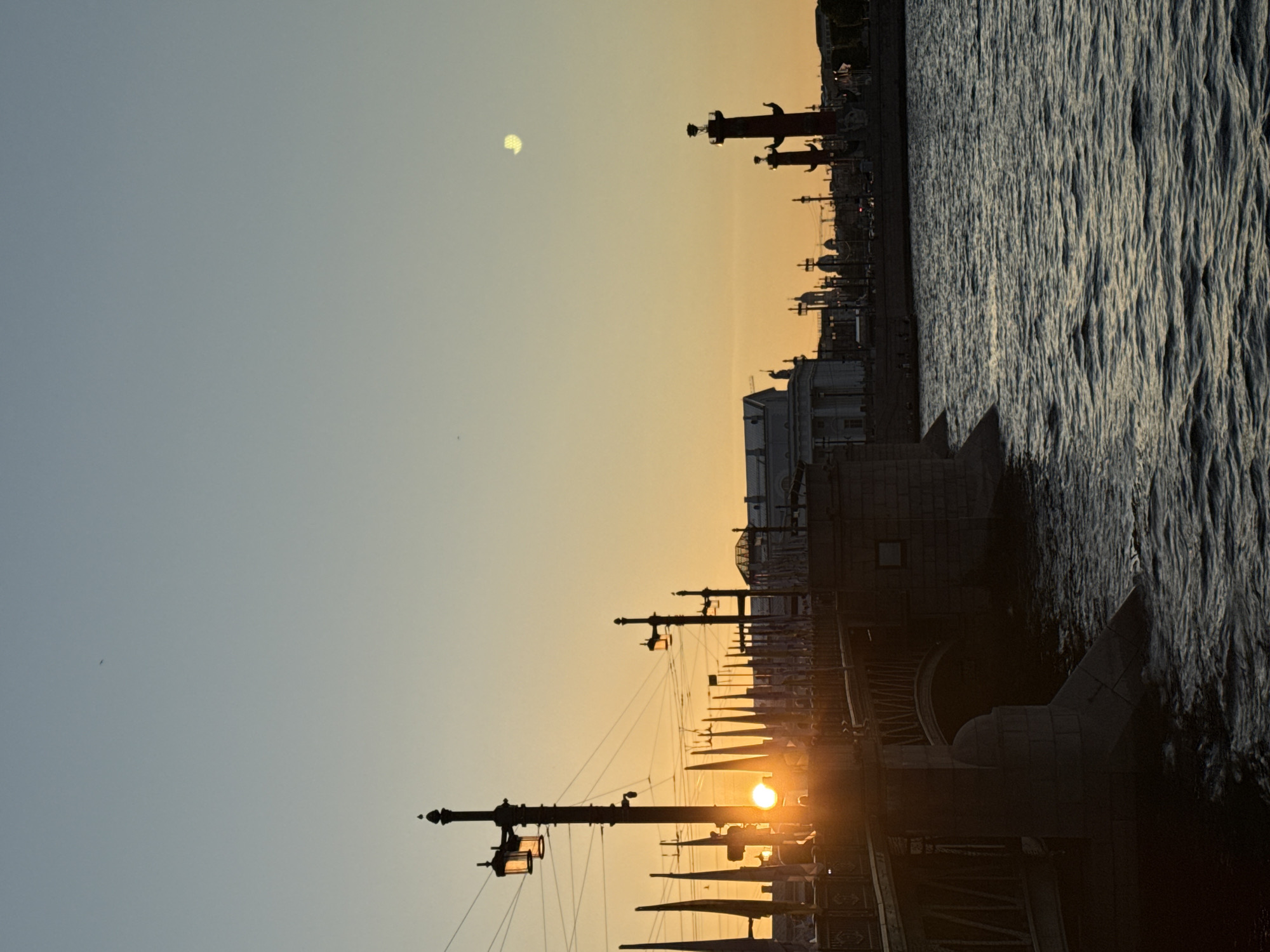 The image presents a panoramic view of the Neva River in Saint Petersburg, Russia, observed during either sunrise or sunset. The scene is heavily backlit, creating strong silhouettes of the architectural and urban elements against a vibrant sky.
In the foreground, to the left, a stone bridge, identifiable as the Palace Bridge, spans the river. The sun, a bright circular disc, is positioned low on the horizon, partially obscured by the bridge's intricate metal railing and an ornate streetlight post, which has its lanterns illuminated. Electrical wires crisscross above the bridge structure. The river's surface directly beneath the bridge exhibits numerous ripples and subtle waves.
The mid-ground is dominated by the wide expanse of the Neva River, with its water displaying a textured pattern of small, reflective waves.
The background reveals the prominent skyline of Vasilyevsky Island. On the right, two iconic red Rostral Columns stand distinctly silhouetted against the warm sky. To their left, a sequence of classical buildings includes a large neoclassical structure, recognized as the Old Saint Petersburg Stock Exchange (Birzha), along with other domes and architectural features of the city.
The sky transitions from a pale blue at the top to warm shades of orange and yellow near the horizon. A distinct crescent-shaped object, consistent with the moon, is visible in the upper-right portion of the sky. Several small, dark specks, likely birds or distant aircraft, are present in the upper atmosphere. No specific individuals or explicit human actions are discernible within the frame.